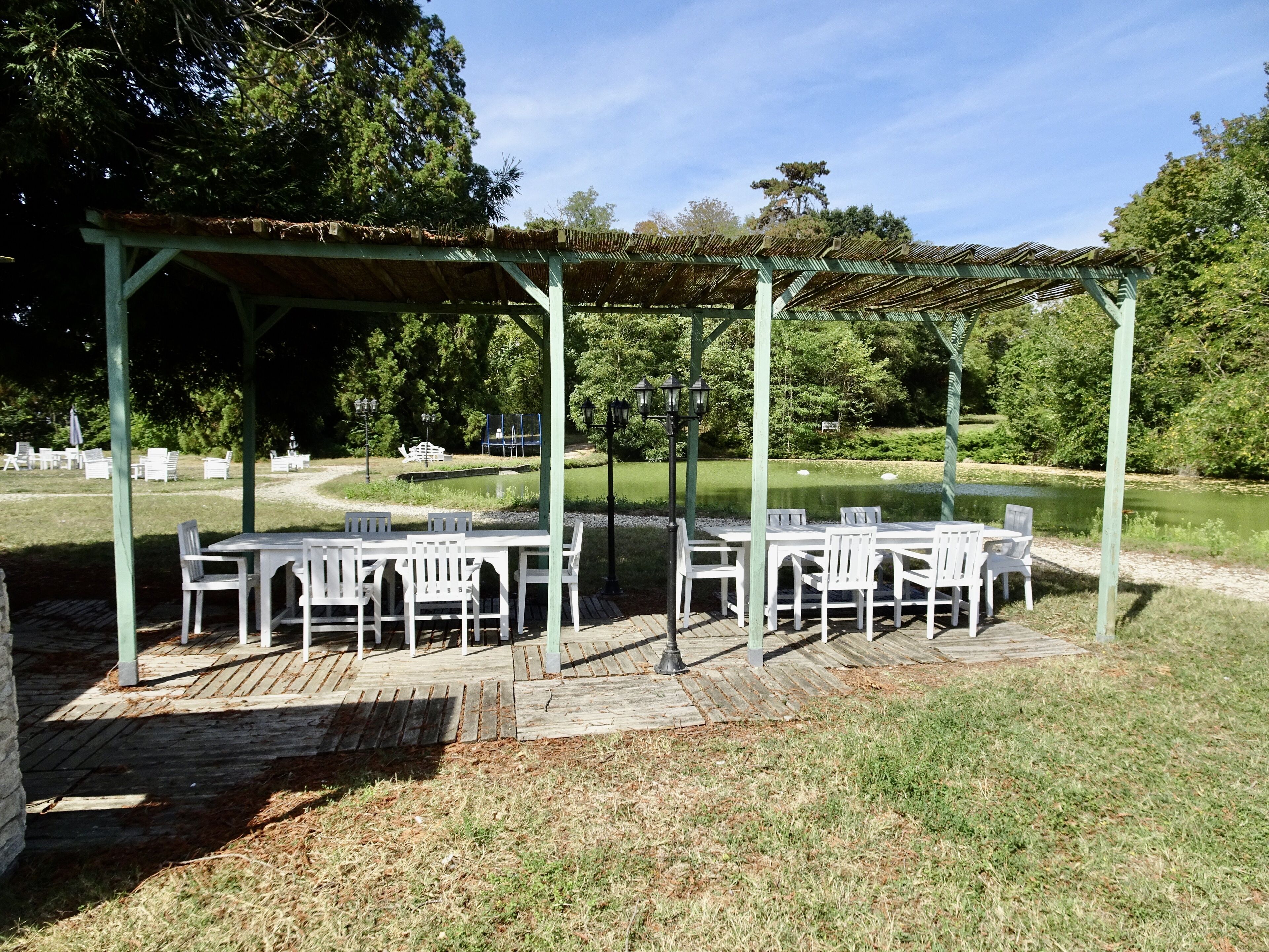 Un patio al aire libre con mesas y sillas blancas, un área de estar cubierta y un estanque al fondo.