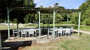 Un patio al aire libre con mesas y sillas blancas, un área de estar cubierta y un estanque al fondo.