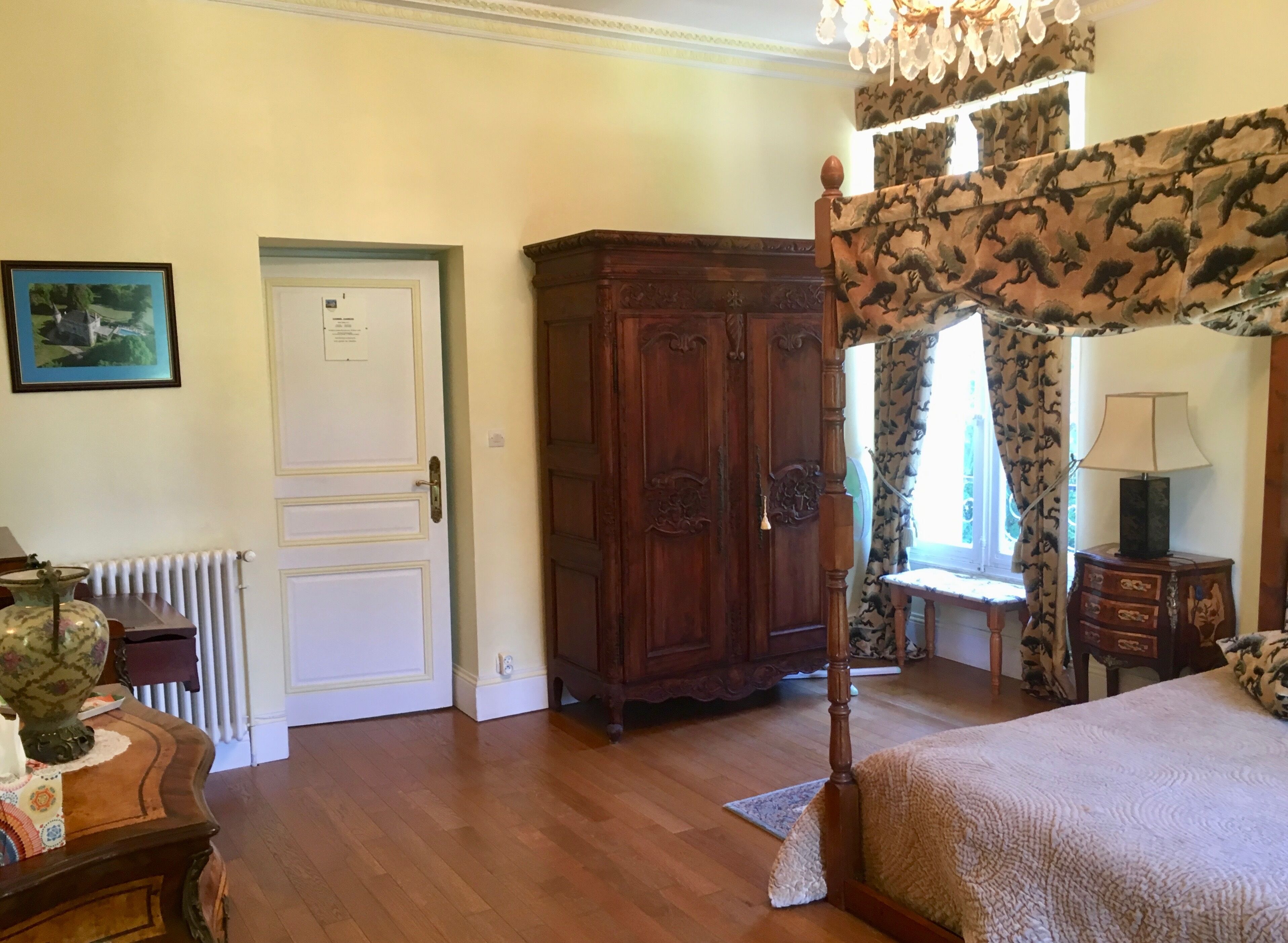 Un dormitorio con cama con dosel, un armario de madera, un radiador, un cuadro enmarcado en la pared y una ventana con cortinas estampadas.