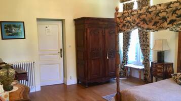Un dormitorio con cama con dosel, un armario de madera, un radiador, un cuadro enmarcado en la pared y una ventana con cortinas estampadas.