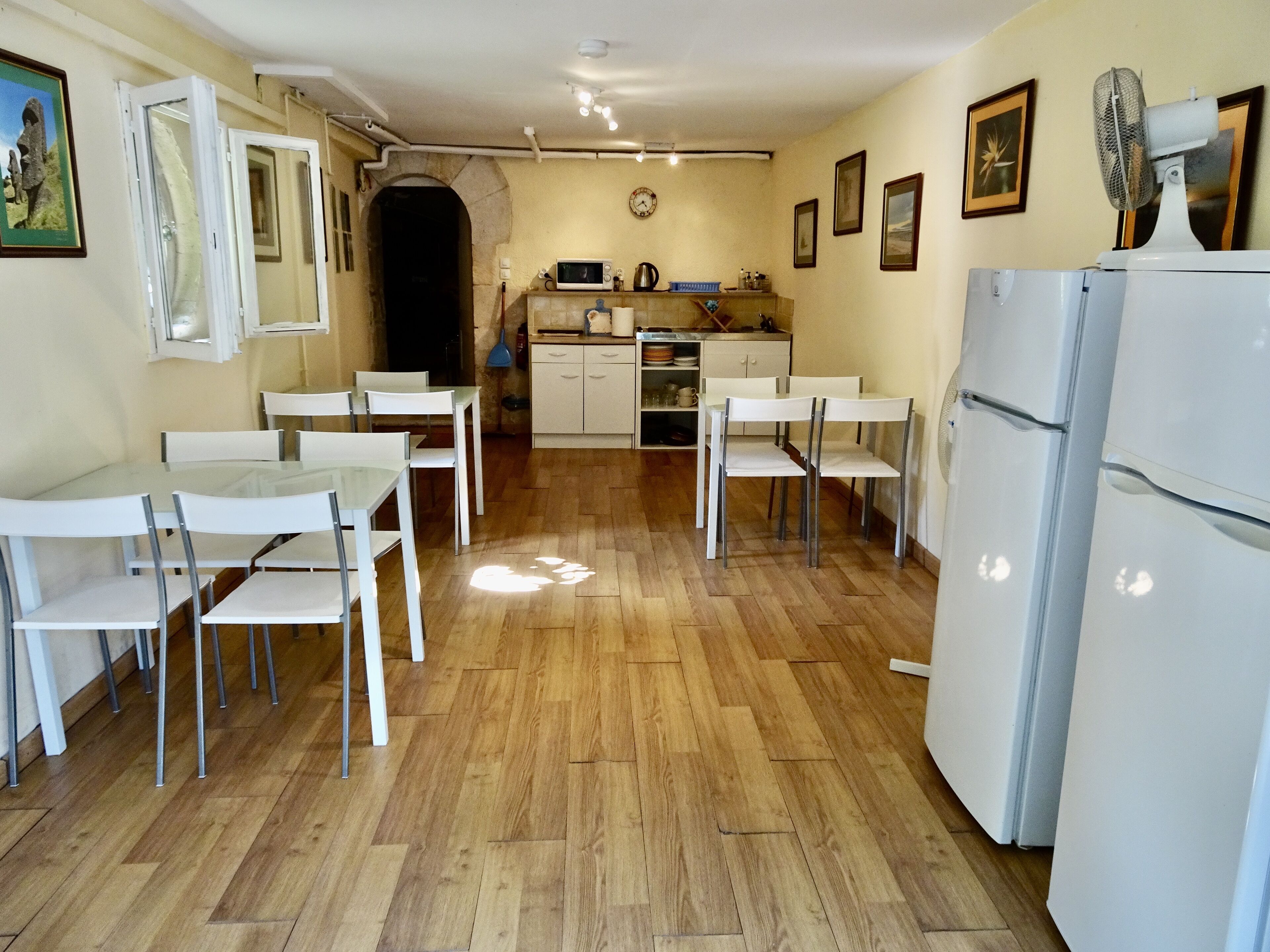 Cocina compacta con armarios blancos, microondas y refrigerador. Hay sillas blancas alrededor de una mesa, y un reloj en la pared.