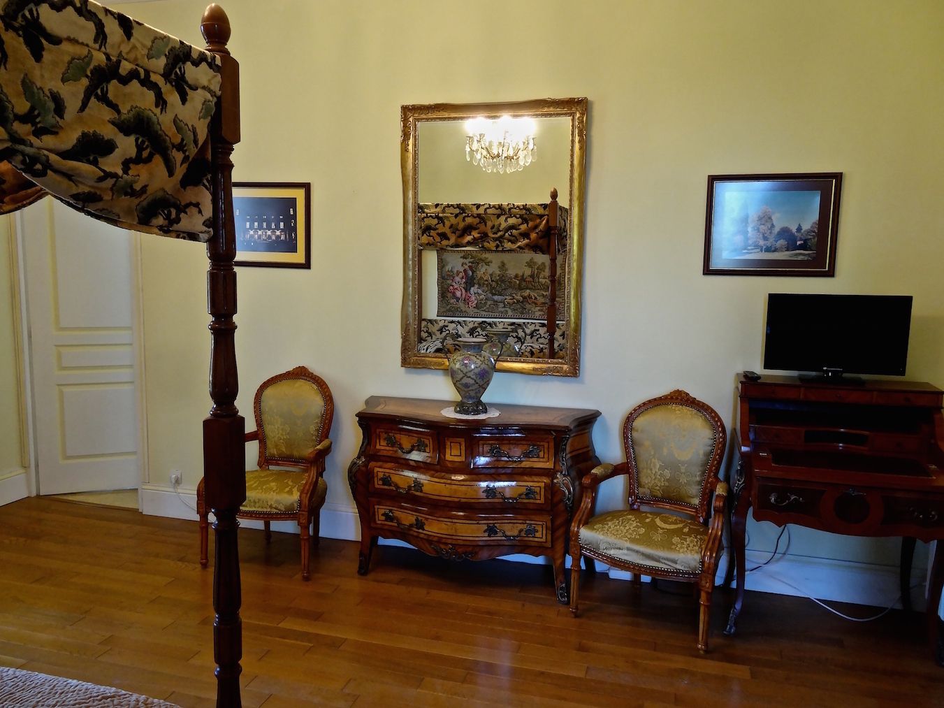 A room with a wooden floor, a bed with a canopy, a television, and two chairs.