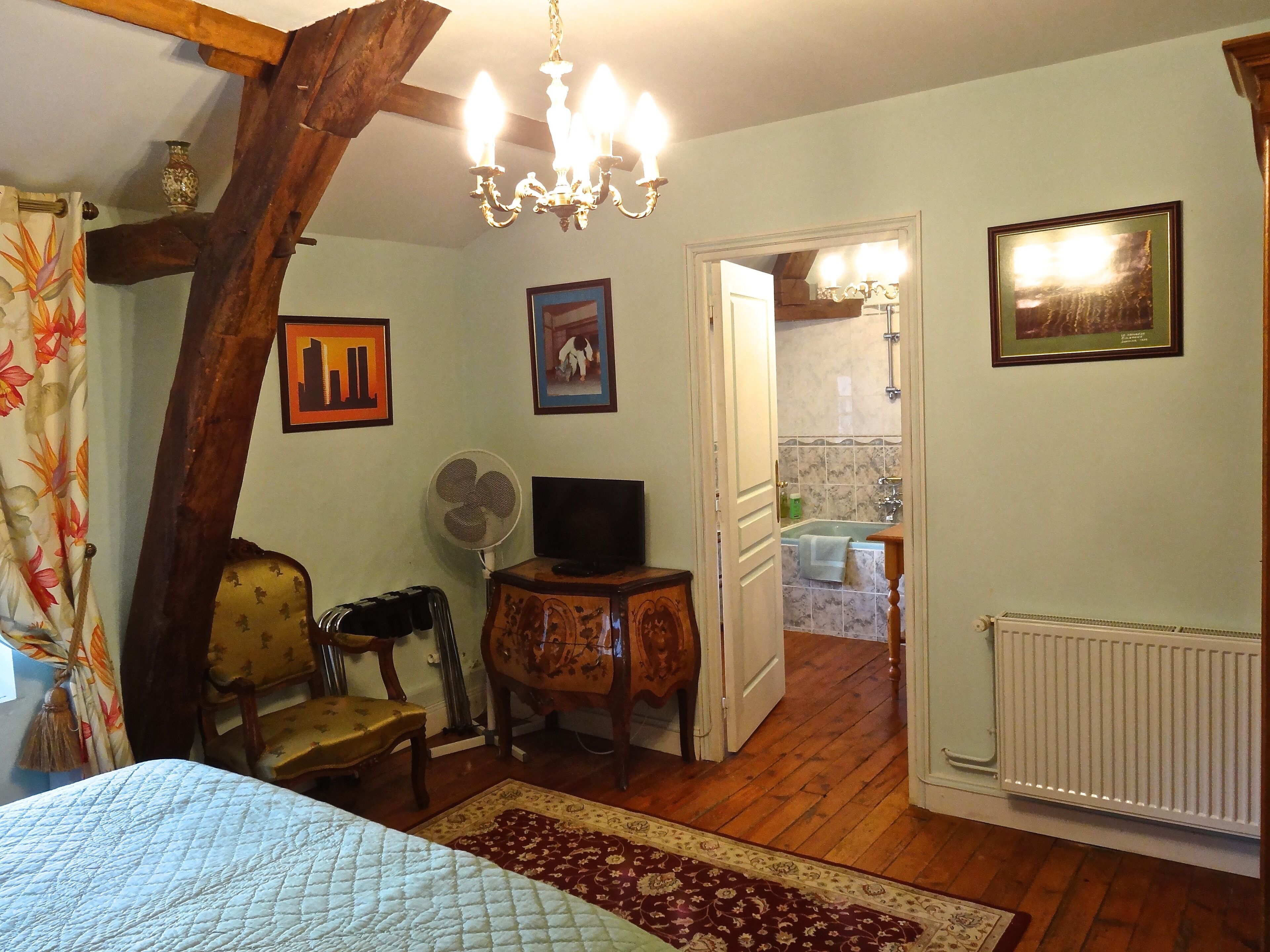 A bedroom with a bed, a wooden chair, a small table with a television, a fan, and a radiator.