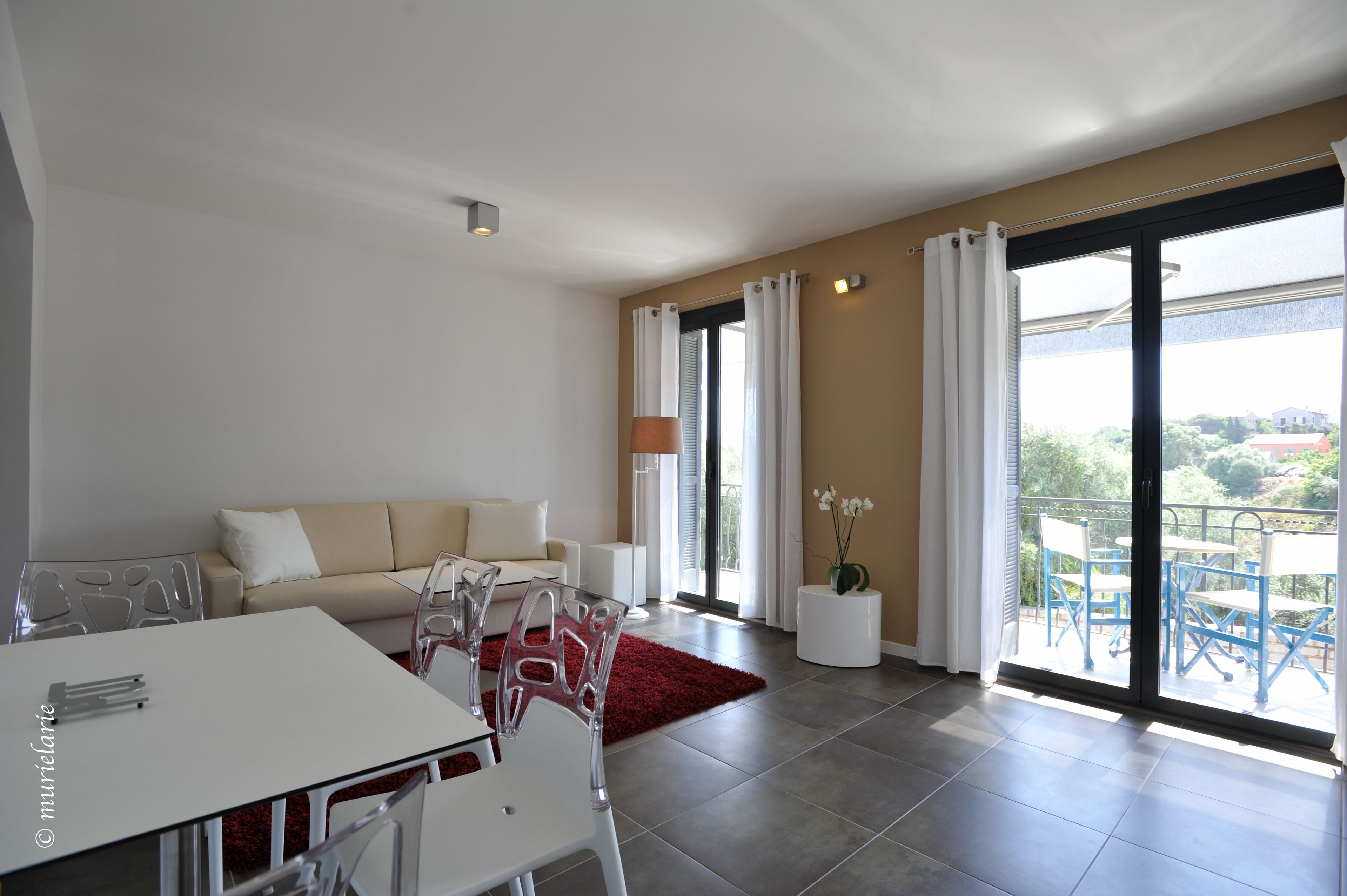 A modern living room with a sofa, dining table, and chairs. There is a large sliding glass door leading to a balcony with outdoor furniture.