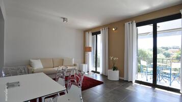 A modern living room with a sofa, dining table, and chairs. There is a large sliding glass door leading to a balcony with outdoor furniture.
