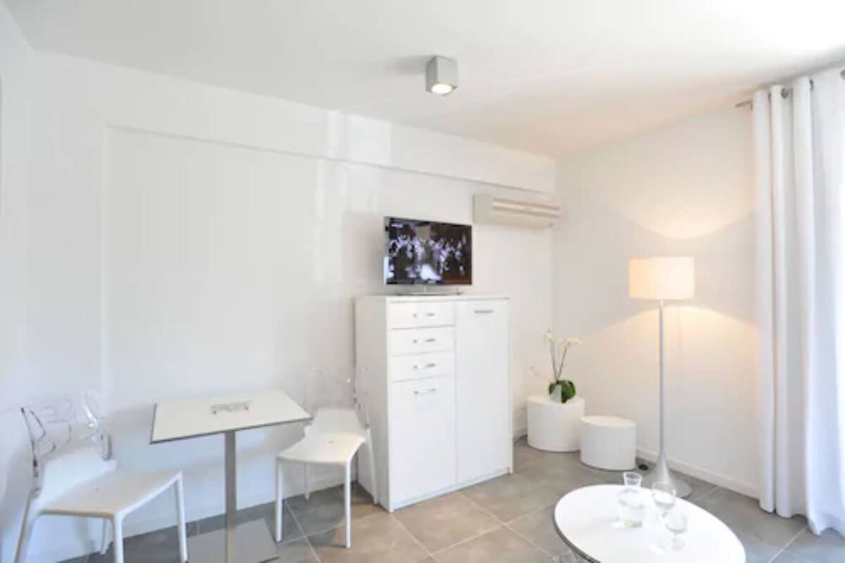 A modern, minimalistic hotel room with a flat-screen TV mounted on the wall, a small desk with a chair, a white cabinet, a round table with glasses, and a floor lamp.