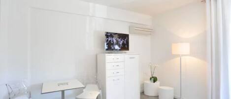 A modern, minimalistic hotel room with a flat-screen TV mounted on the wall, a small desk with a chair, a white cabinet, a round table with glasses, and a floor lamp.