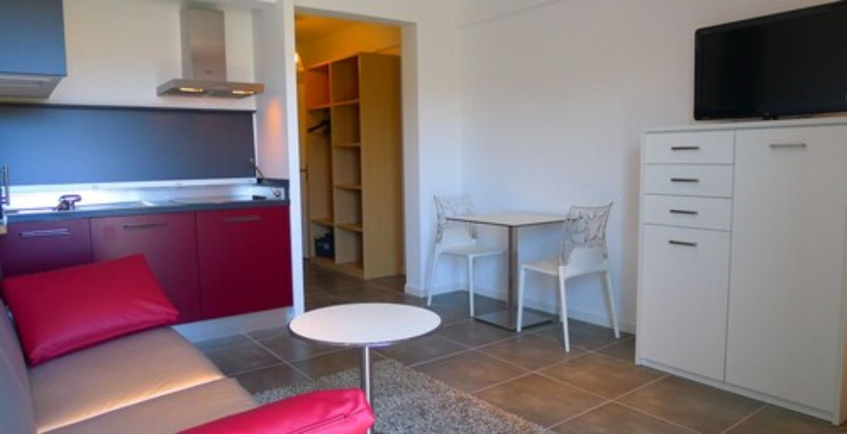 Um espaço compacto com cozinha americana, área de jantar com duas cadeiras, mesa redonda pequena e TV fixada na parede.