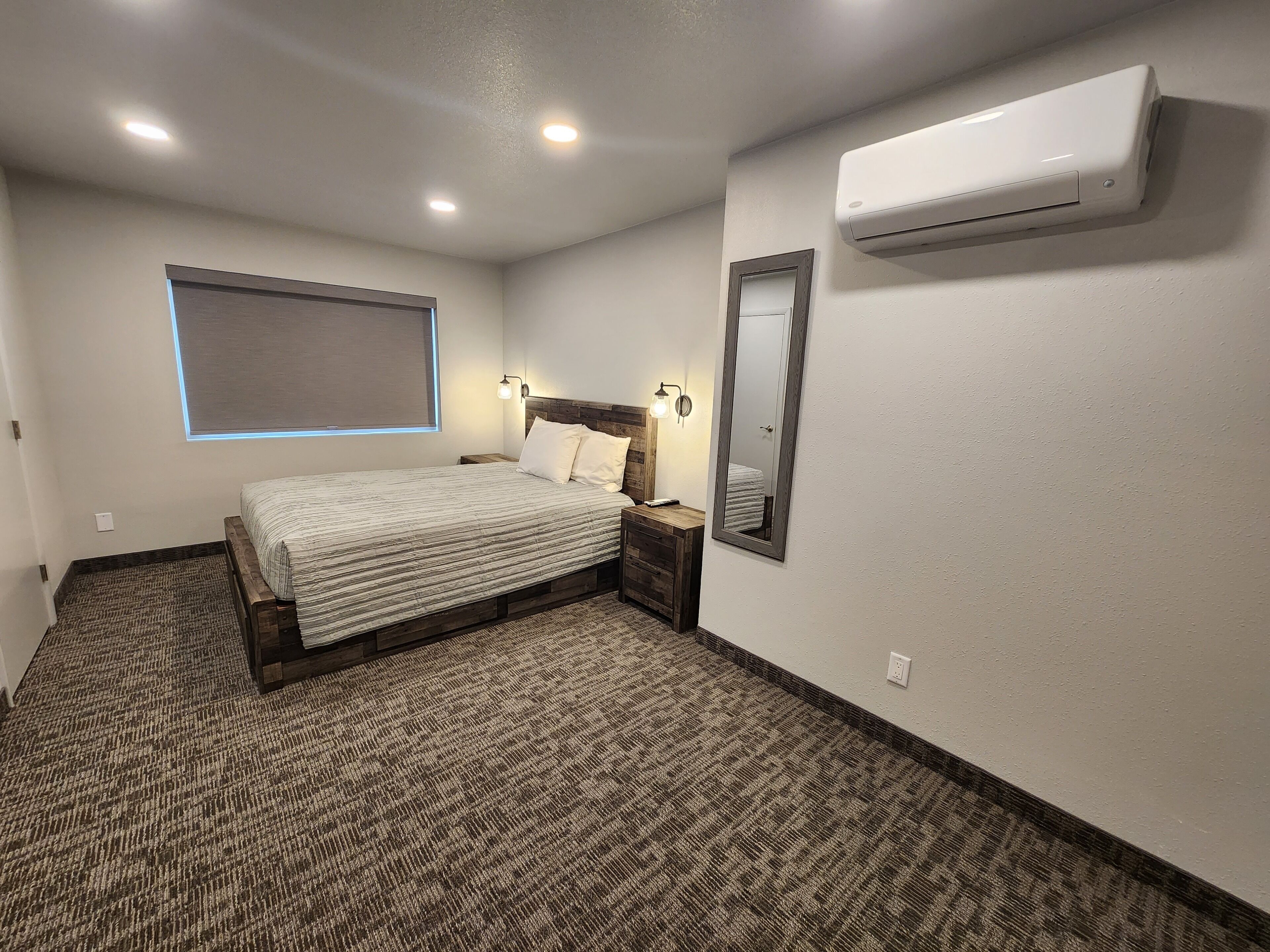 A bedroom with a bed, bedside tables, a wall-mounted air conditioner, a window with blinds, and a carpeted floor.