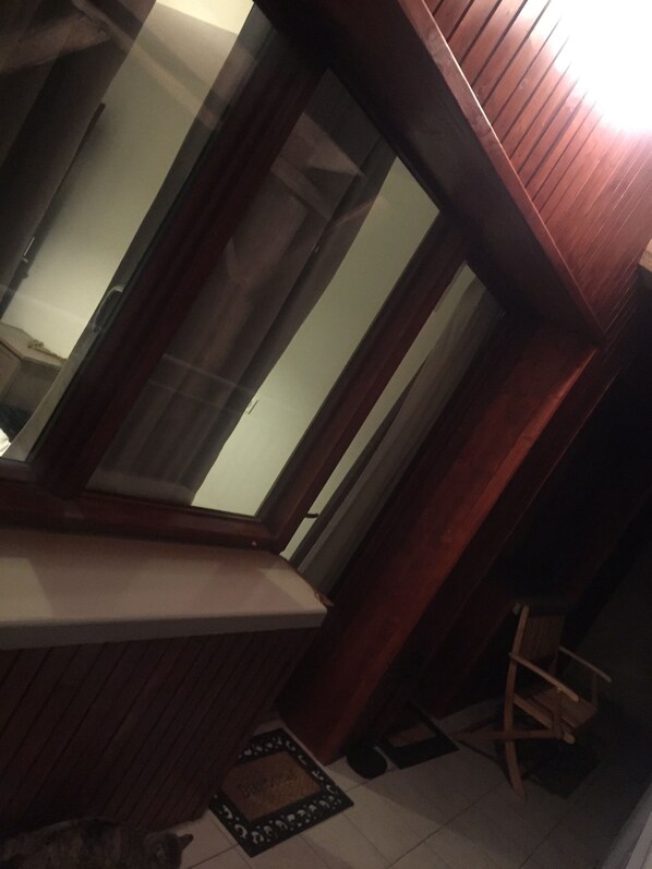 A room with a wooden bench, a small table, and a chair. There is a window with blinds, a door, and a doormat on the floor.