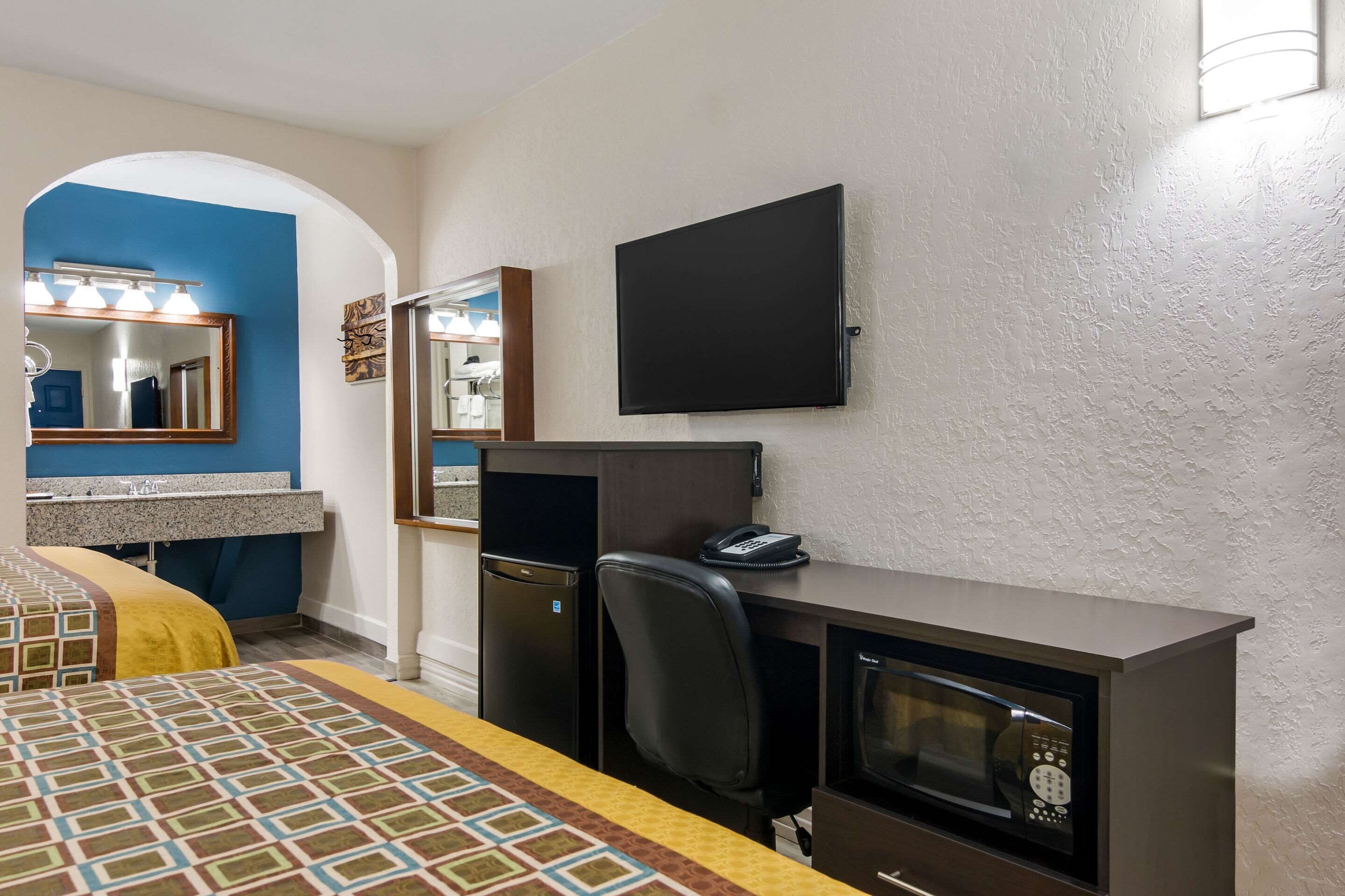 Ein Hotelzimmer mit einem Bett, einem Schreibtisch mit Computer, einer Mikrowelle, einem Kühlschrank, einem Fernseher und einem Badezimmer mit Spiegel und Waschbecken.