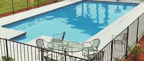 Una piscina rectangular con valla metálica, rodeada de acolchado rojo y equipada con mobiliario exterior.