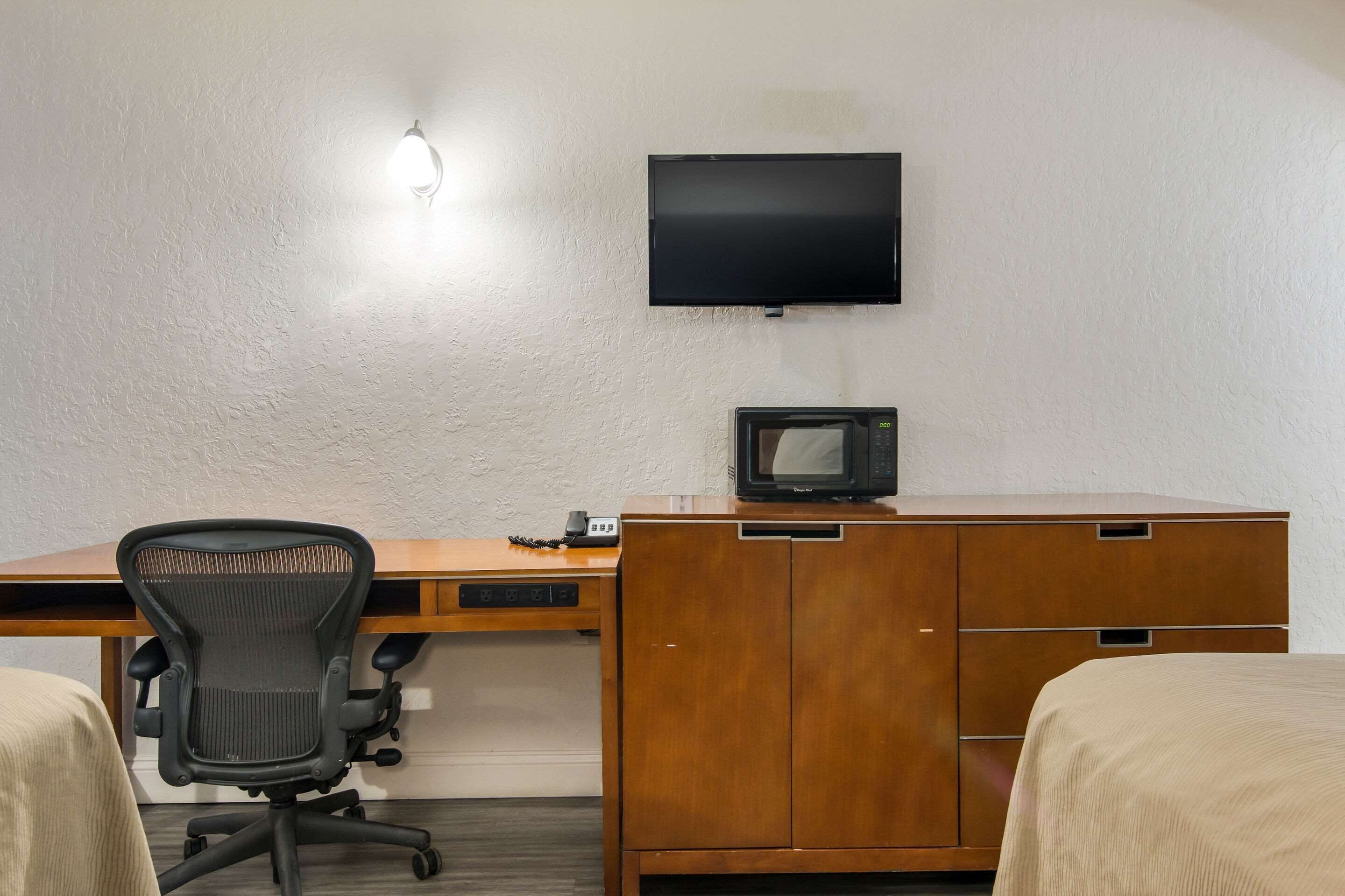 A hotel room with a wooden desk, a black office chair, a television mounted on the wall, a microwave, and two beds.