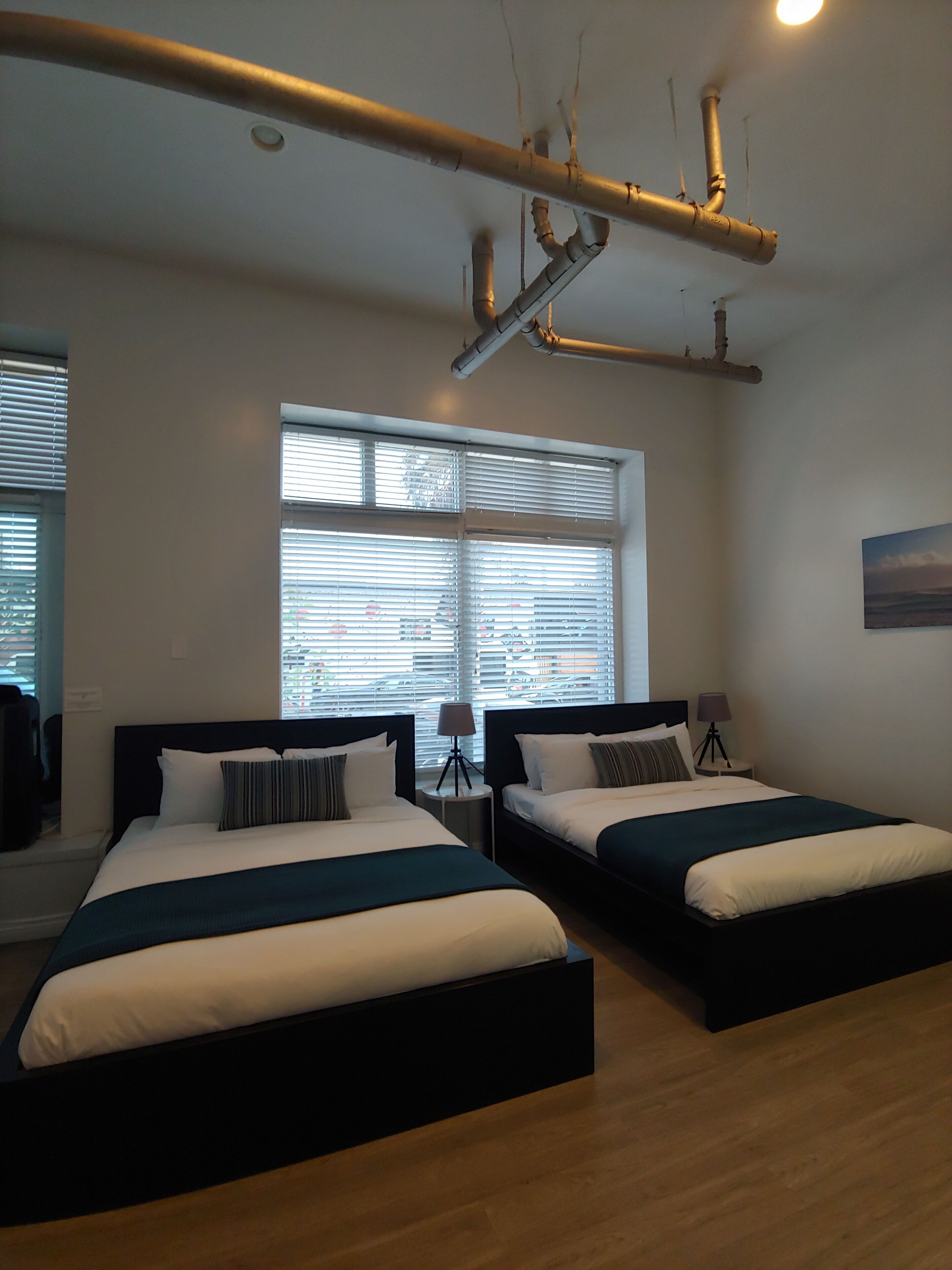 Una habitación de hotel con dos camas, un ventanal con persianas, una lámpara sobre una mesita de noche y conductos expuestos en el techo.