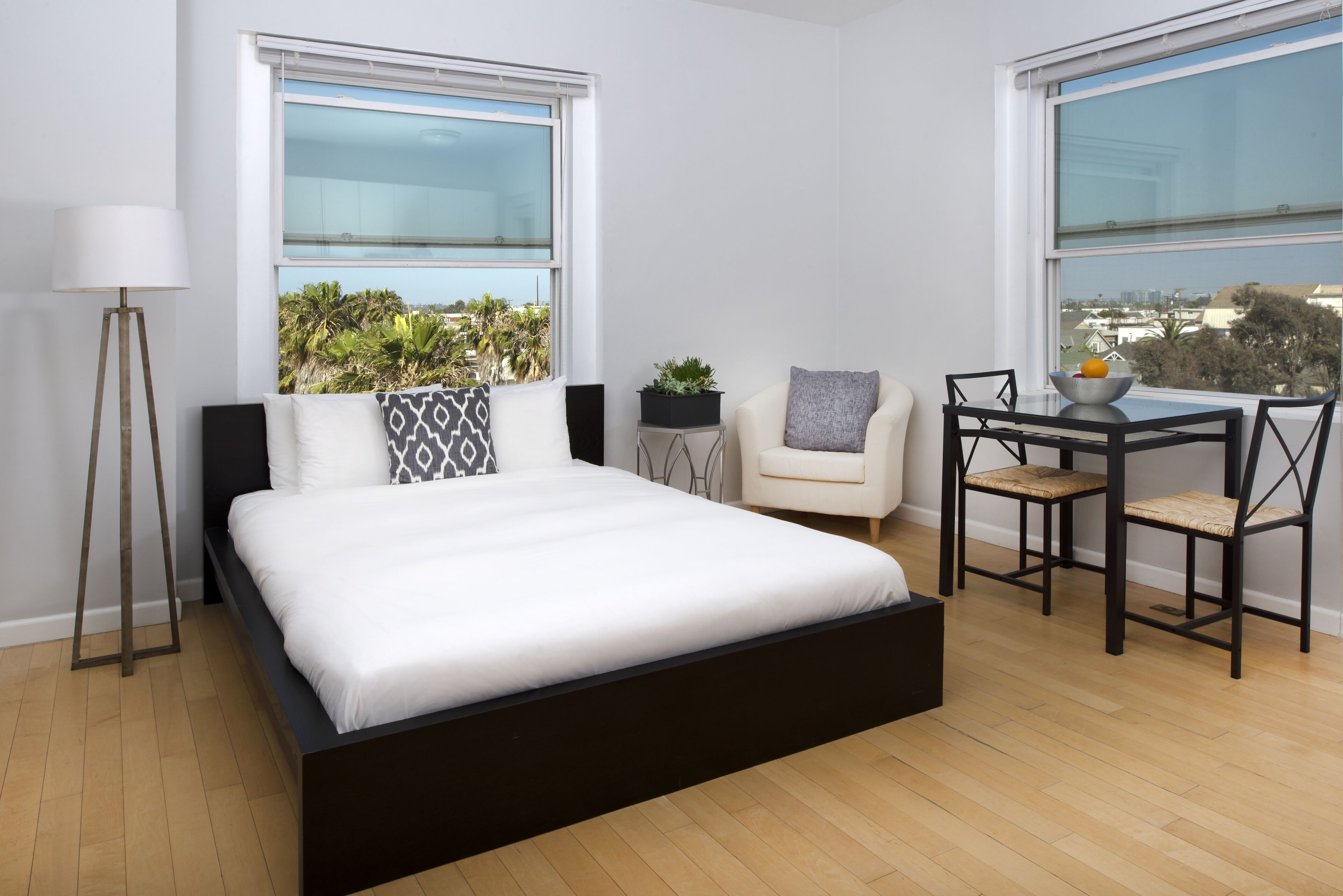 Un dormitorio moderno con una cama grande, dos ventanas, una mesita de noche, una silla y una pequeña mesa con un frutero.