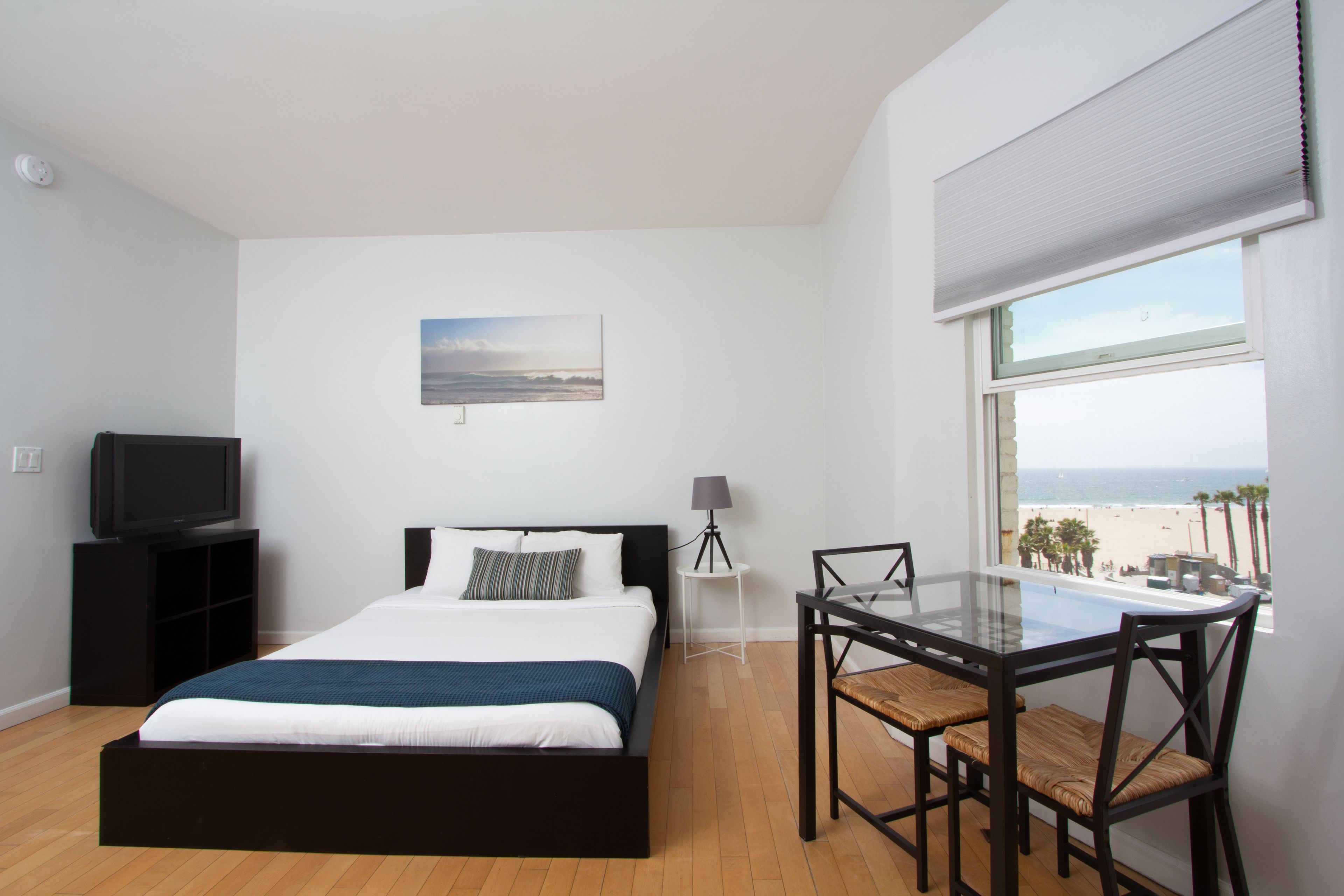 Quarto com cama, televisão, escrivaninha com cadeiras, abajur e janela com vista para a praia.