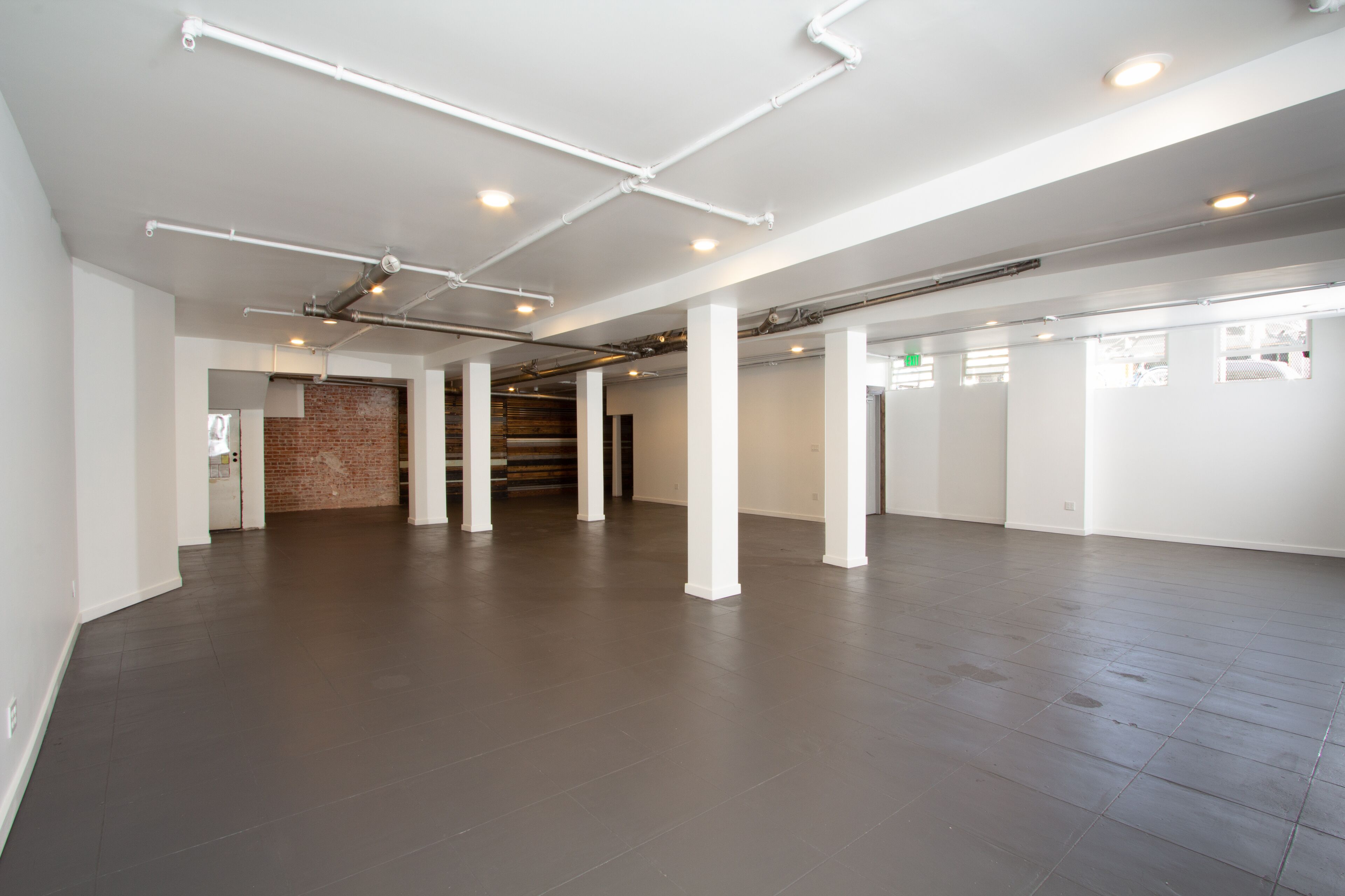A spacious, empty room with a dark floor, white walls, and exposed ductwork on the ceiling.
