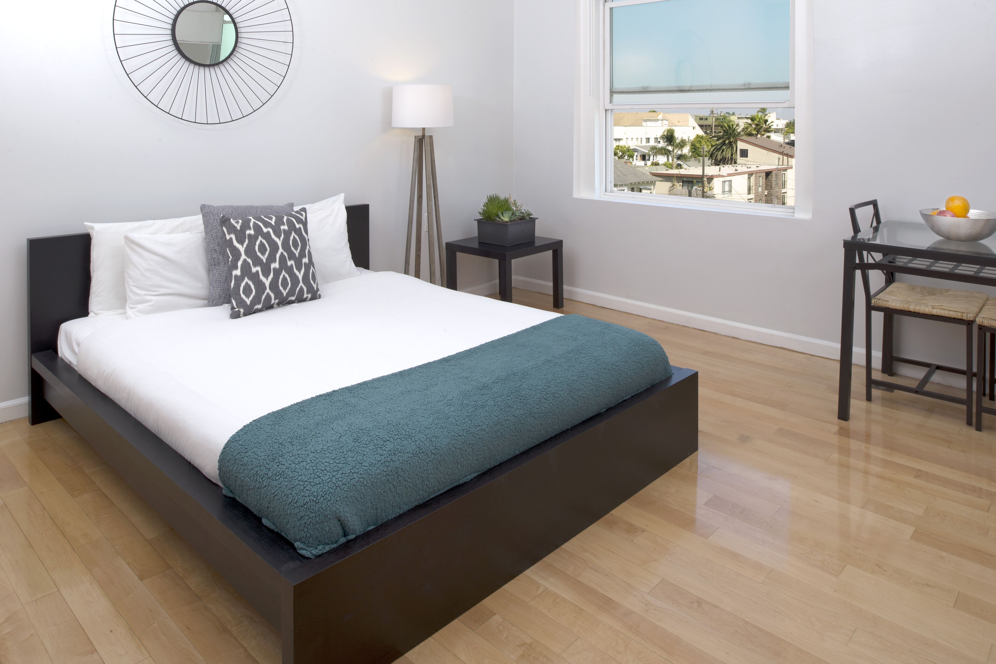 A modern bedroom with a large bed, a bedside table with a lamp, a small plant, and a view of the city through a window.
