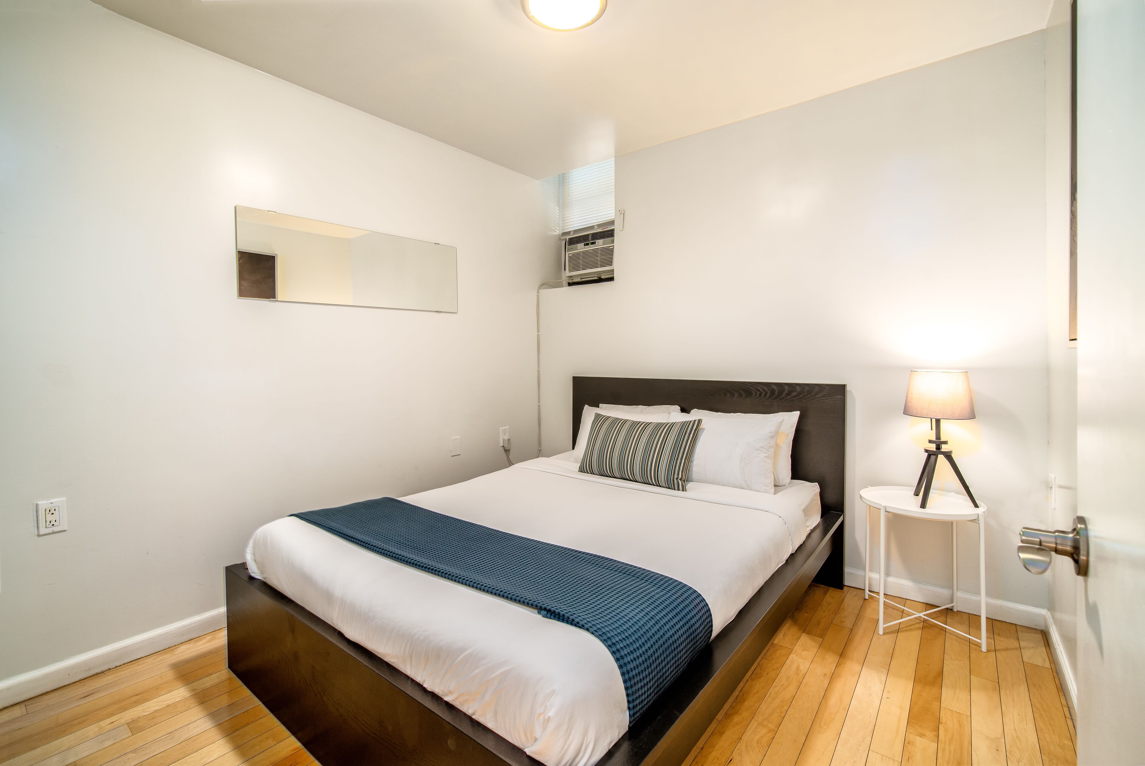 A modern bedroom with a large bed, a bedside lamp, and a wall-mounted air conditioner.