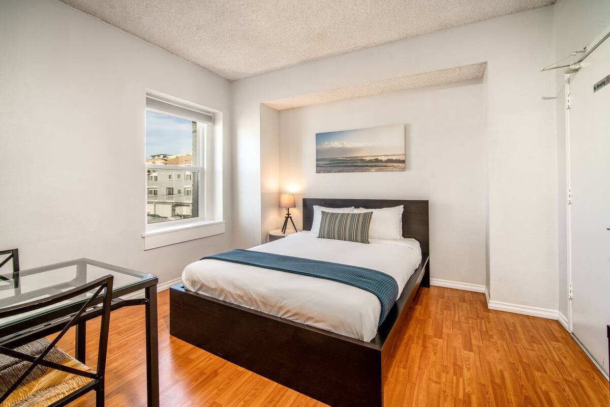 Ein Schlafzimmer mit einem Bett, einem Glastisch, einem Stuhl, einem Fenster und einem Gemälde an der Wand.