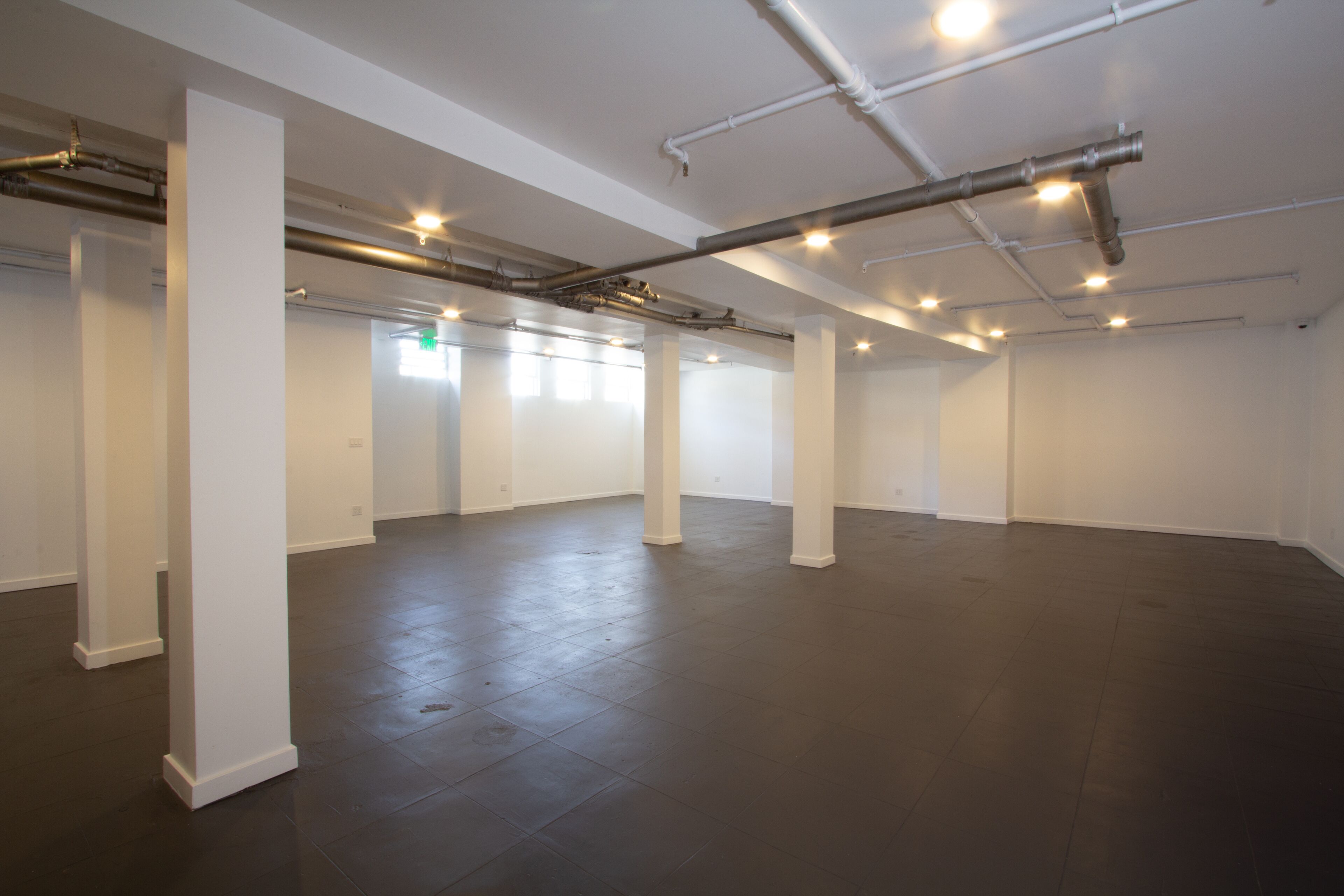 A spacious, empty room with white pillars, dark flooring, and visible ceiling pipes.
