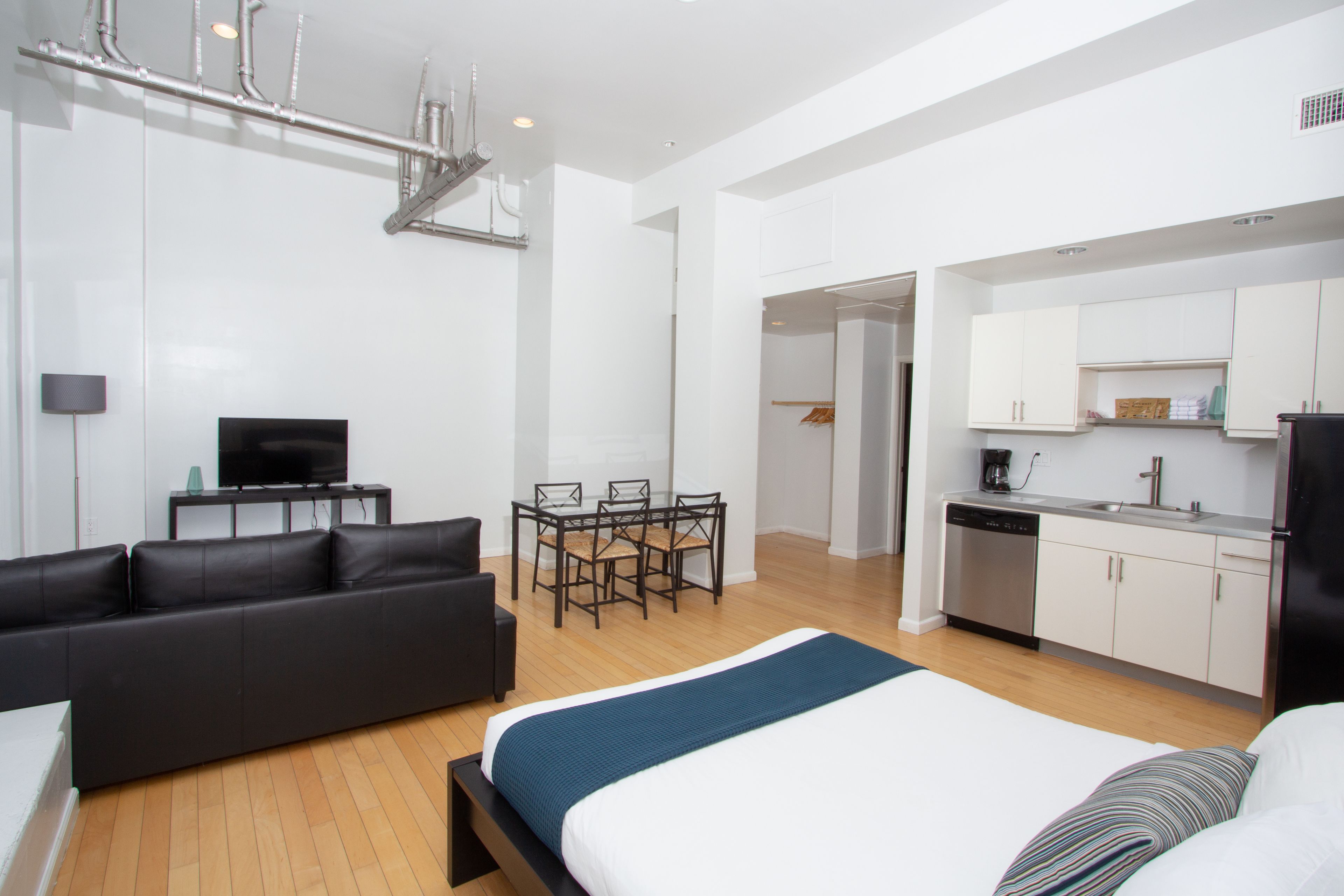 A modern hotel room with a bed, a sofa, a dining table with chairs, a kitchen area, and a TV.