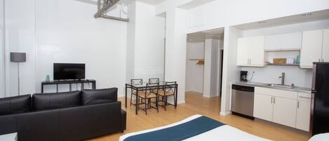 A modern hotel room with a bed, a sofa, a dining table with chairs, a kitchen area, and a TV.