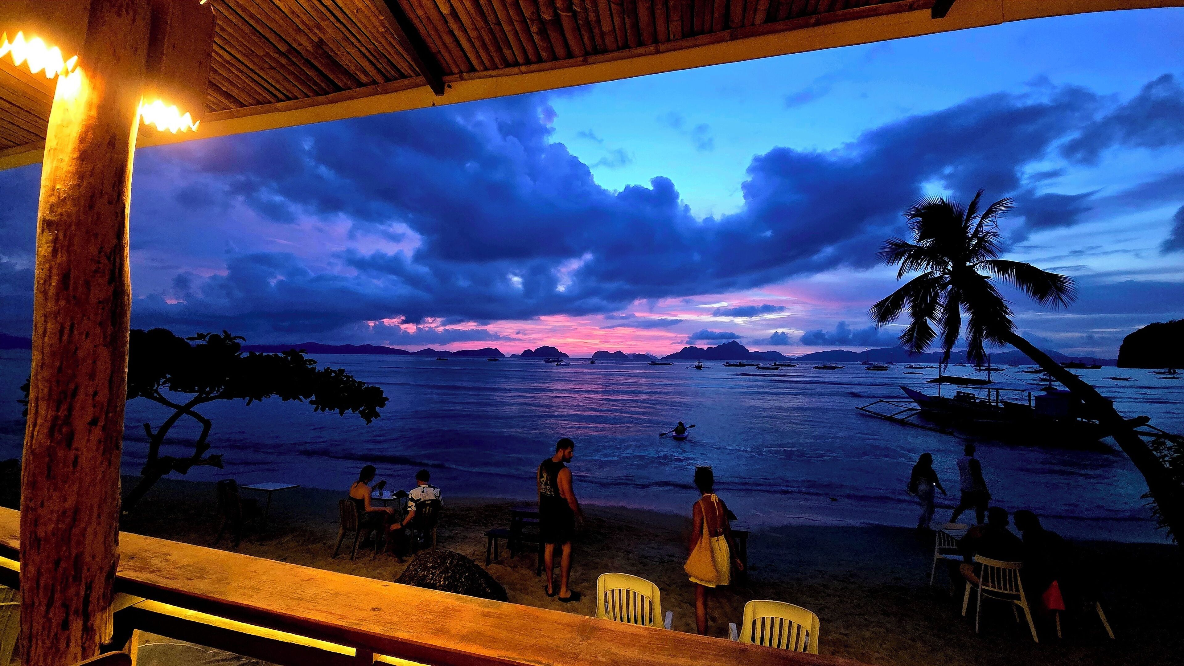 A beach scene with a wooden structure, chairs, and tables under a sunset sky.