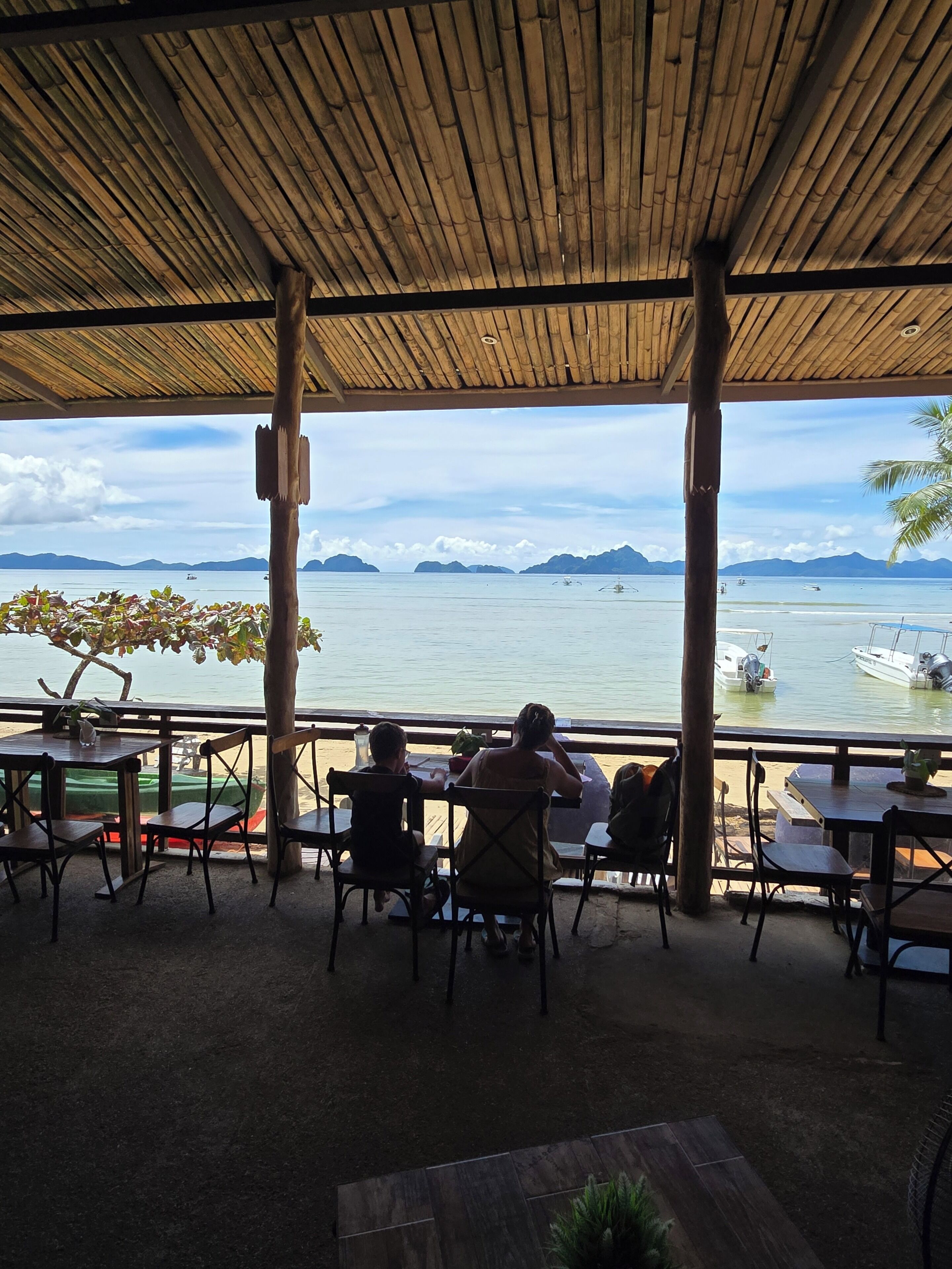 An outdoor seating area with tables and chairs, overlooking a calm sea with boats and distant islands.