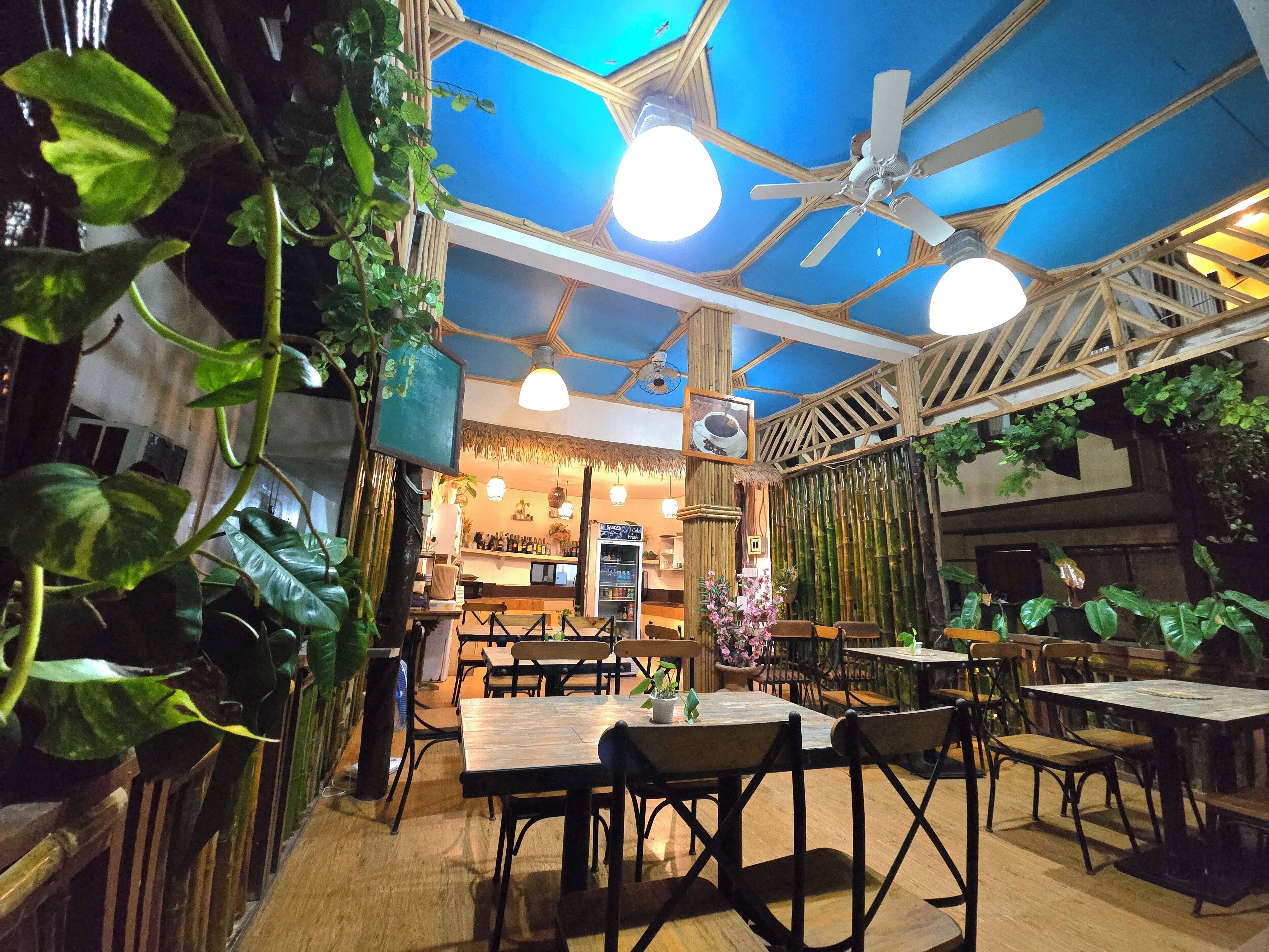A restaurant with wooden tables and chairs, ceiling fans, and hanging plants.