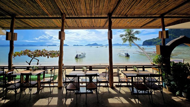 A covered outdoor seating area with tables and chairs, overlooking a calm sea with boats and distant islands. - Islandfront Cottages (El Nido)