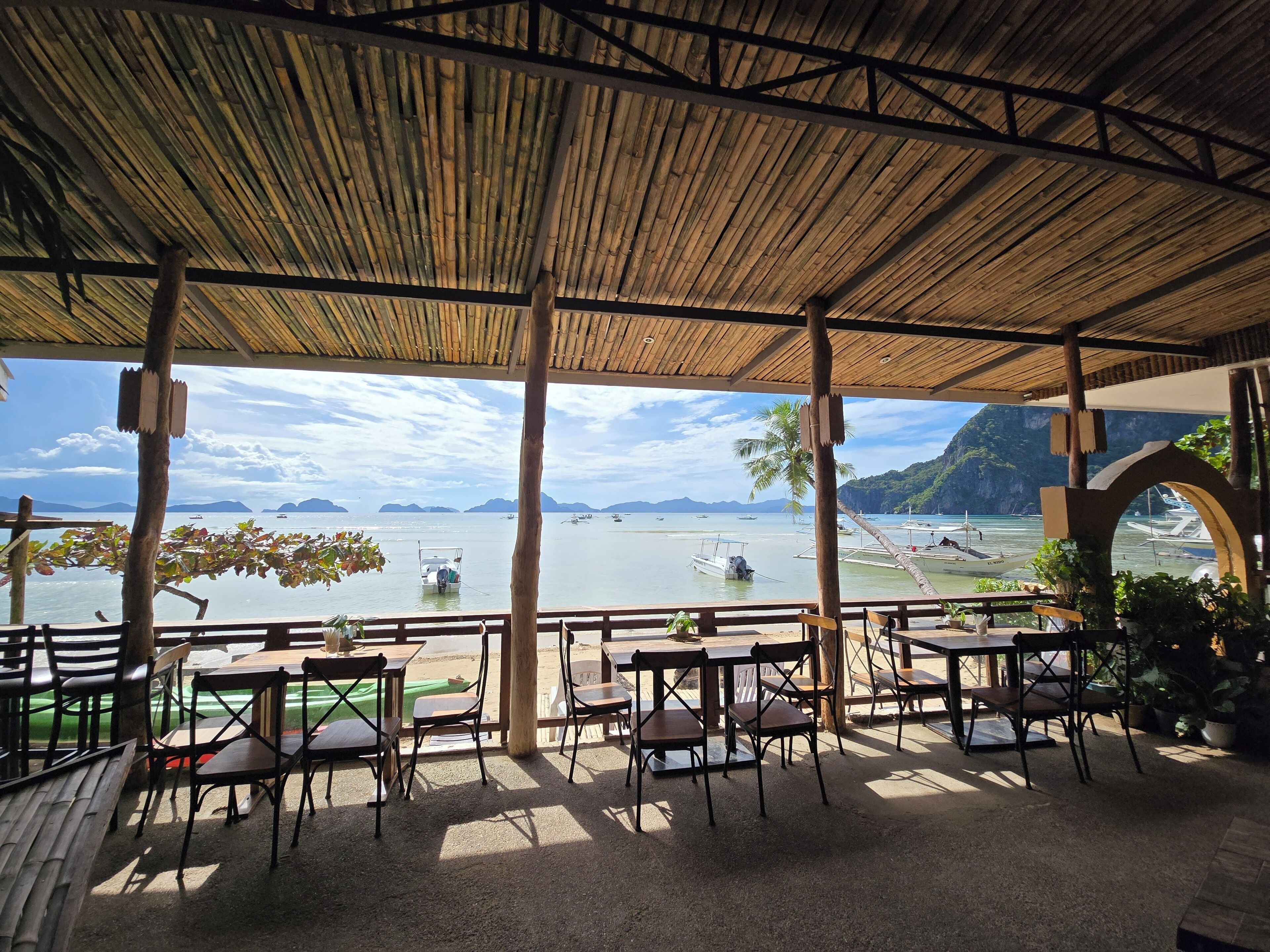 An outdoor seating area with tables and chairs, overlooking a calm body of water with boats and distant islands.