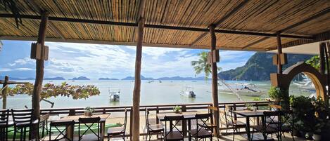 An outdoor seating area with tables and chairs, overlooking a calm body of water with boats and distant islands.