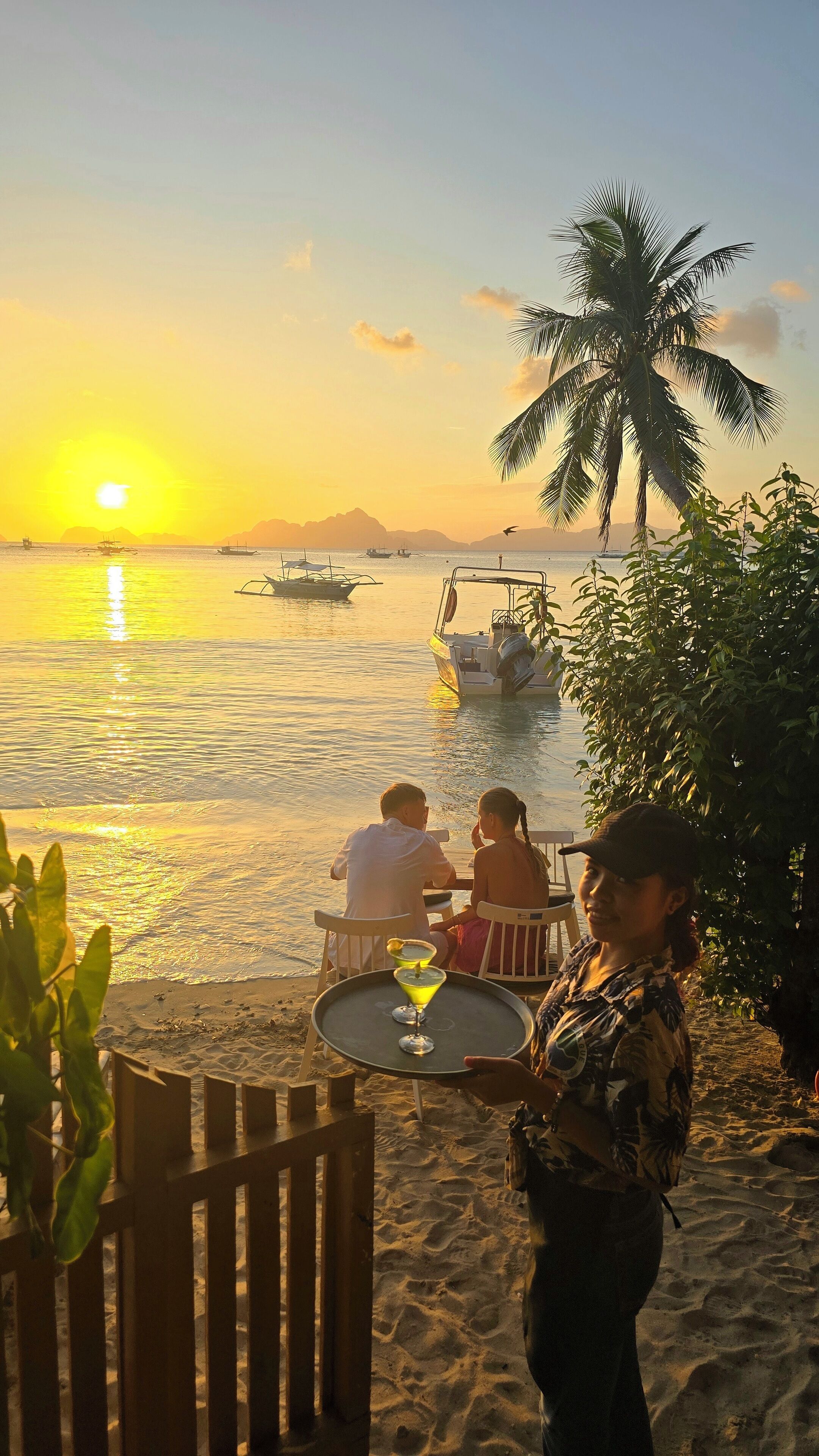 Escena en la playa con atardecer, palmeras y una pareja sentada en una mesa con bebidas.
