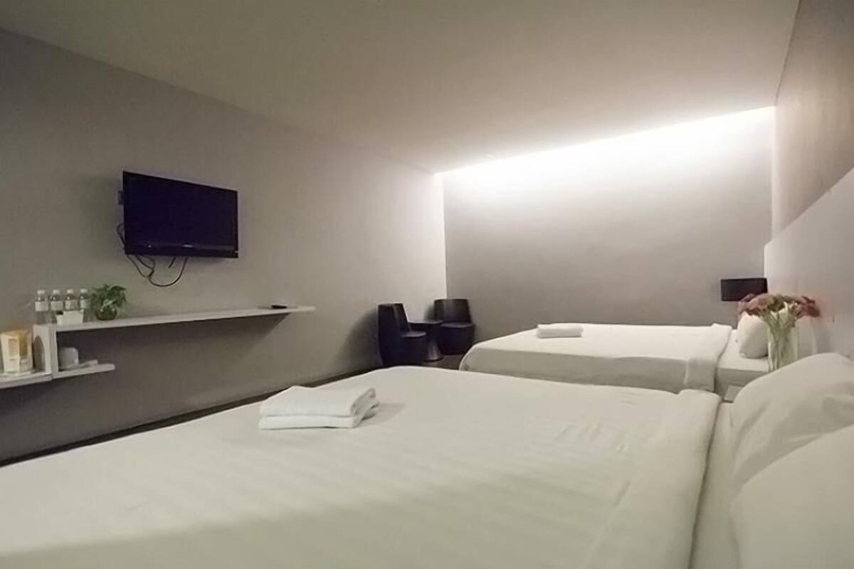 Habitación de hotel con dos camas, un televisor de pantalla plana montado en la pared, una mesita de noche con lámpara y un estante pequeño con artículos de aseo.