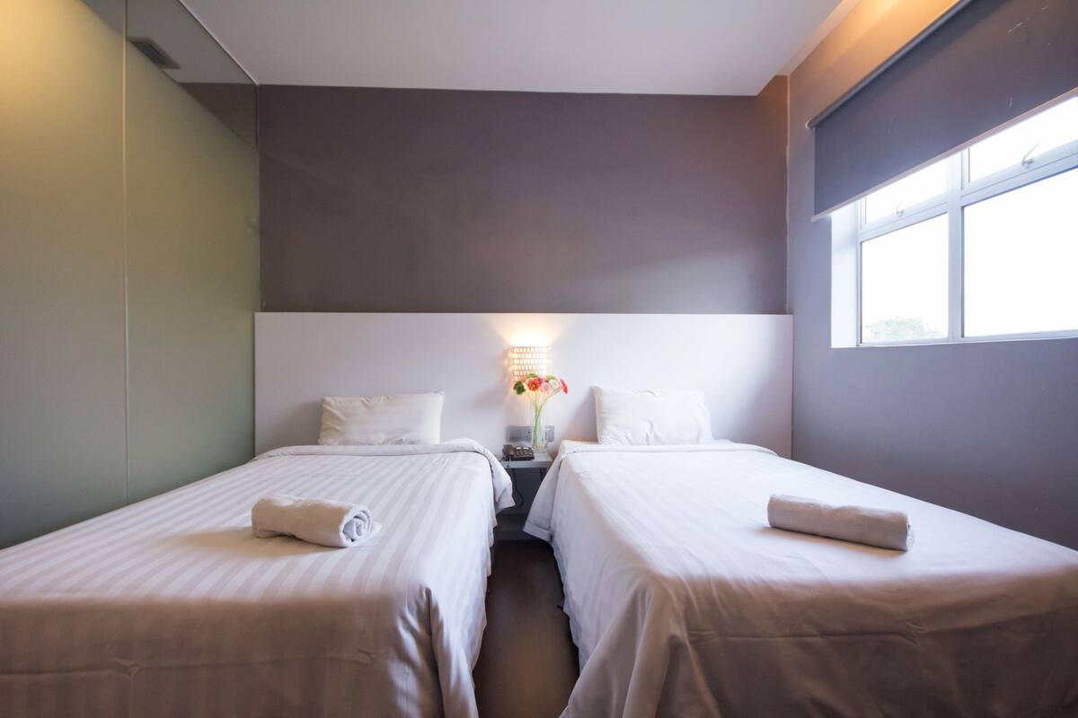 Una habitación de hotel con dos camas individuales, una mesita de noche con lámpara y una ventana.