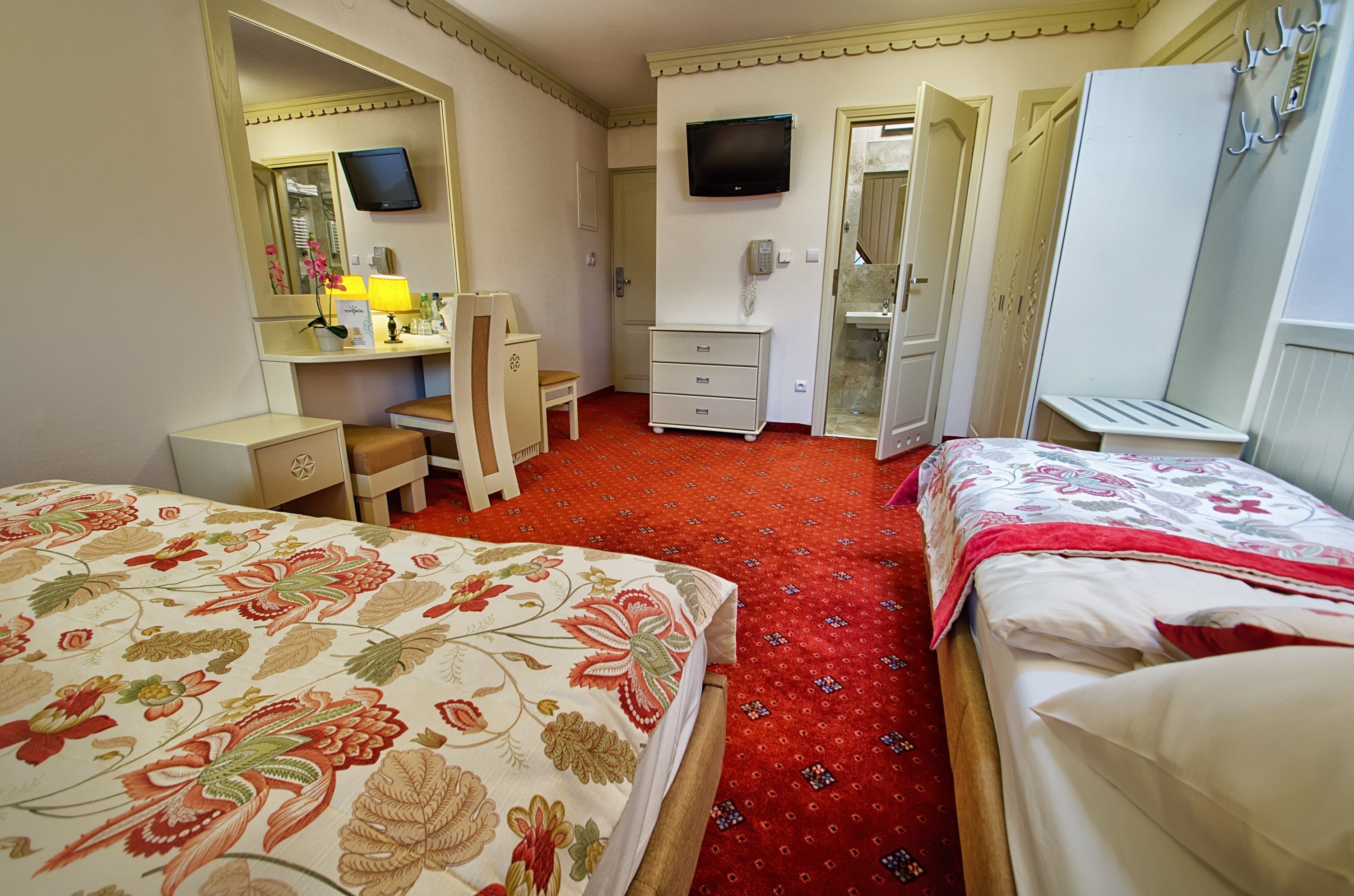 Habitación de hotel con dos camas, un escritorio con silla, espejo, televisor, teléfono y armario.