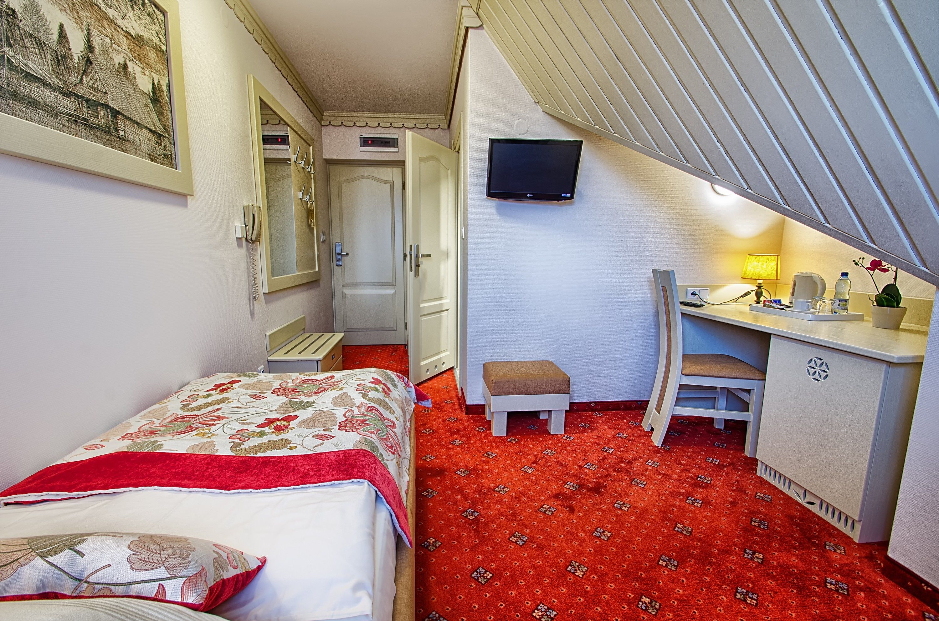 A hotel room with a bed, a desk, a chair, a TV, and a red carpet.