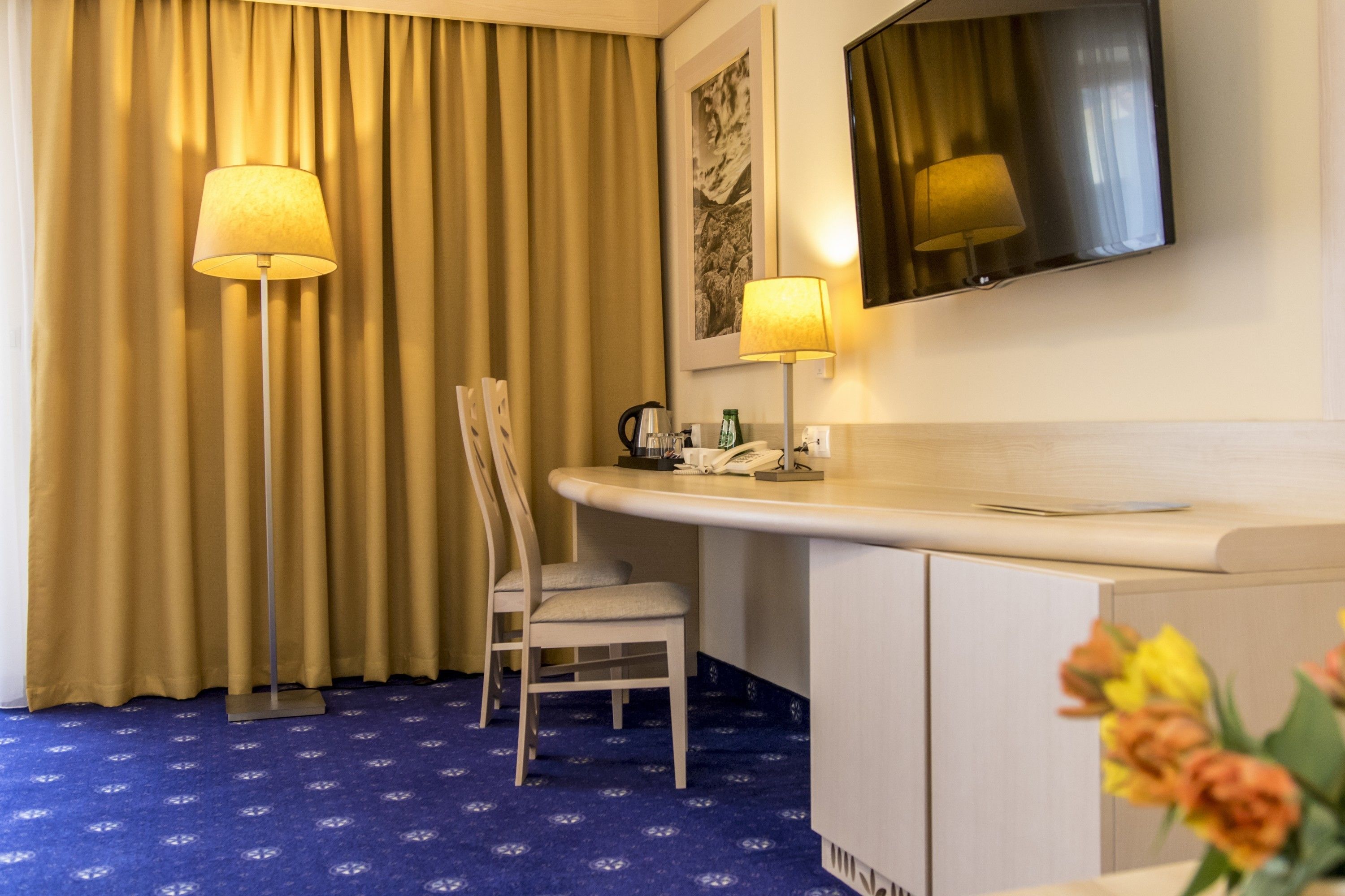A hotel room with a desk, chair, and a large mirror. There is a telephone and a small table with a lamp. The room has a blue carpet and beige curtains.