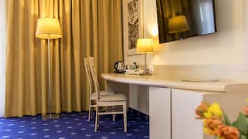 A hotel room with a desk, chair, and a large mirror. There is a telephone and a small table with a lamp. The room has a blue carpet and beige curtains.