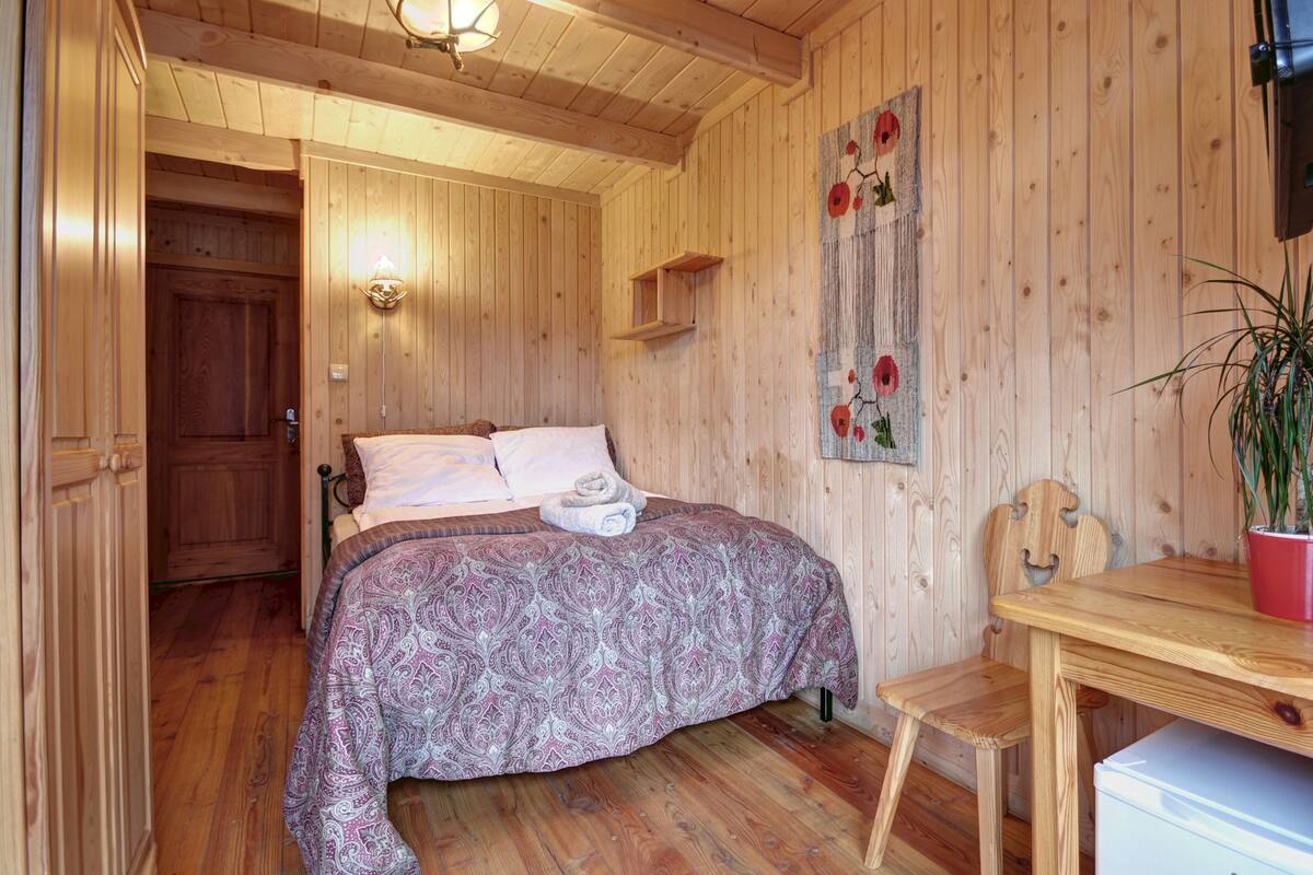 Ein Zimmer mit Holzboden, einem Bett, einem kleinen Tisch, einem Stuhl und einem an der Wand befestigten Regal.