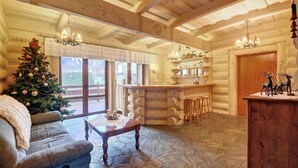 A cozy log cabin interior with a Christmas tree, wooden furniture, and a stone floor.