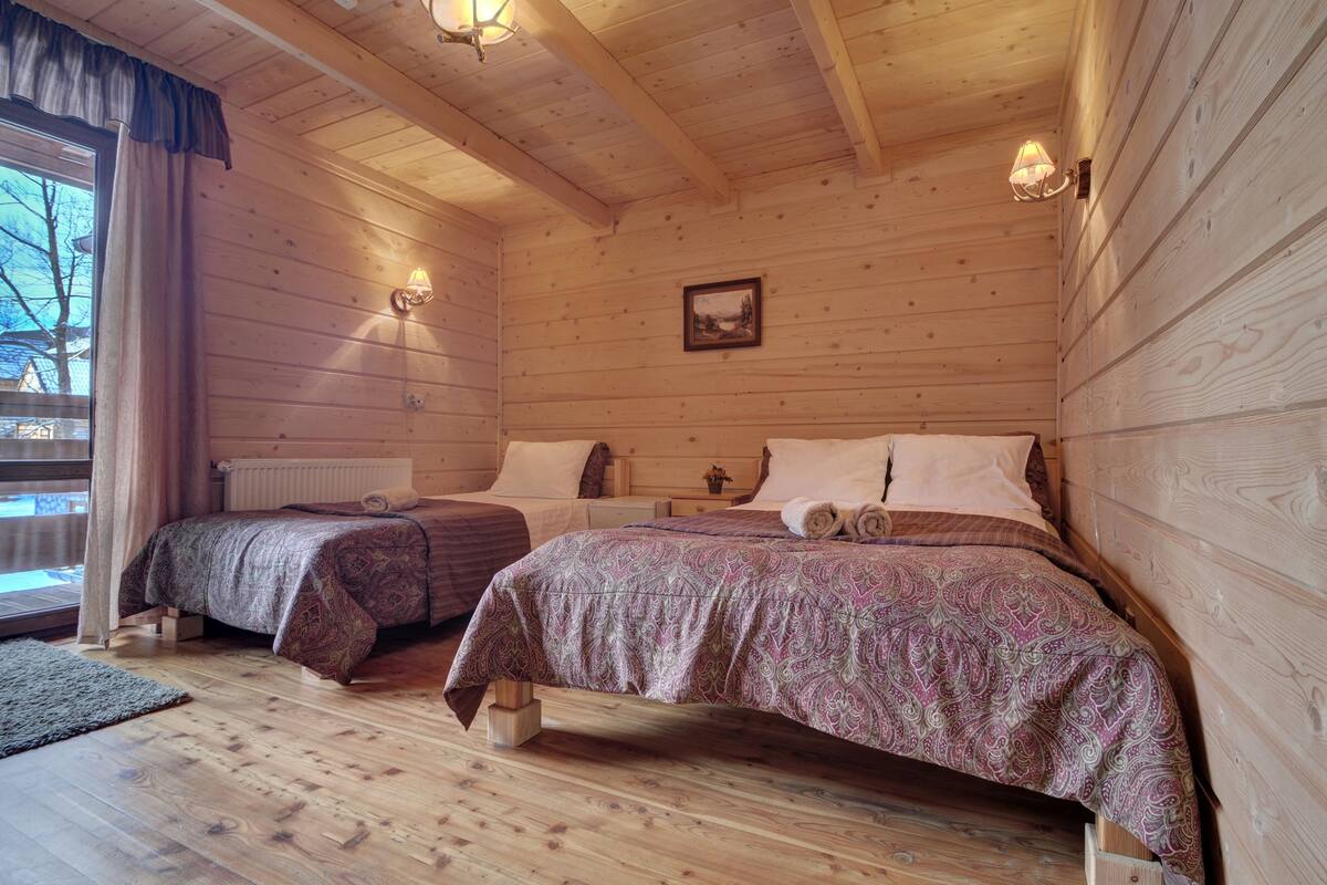 Ein Schlafzimmer einer Holzhütte mit zwei Betten, einem Fenster mit Vorhängen, einem Wandbild und einem Nachttisch.