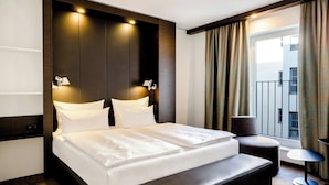 Quarto de hotel moderno com uma cama grande, um sofá pequeno, uma mesa auxiliar e vista para a cidade pela janela.