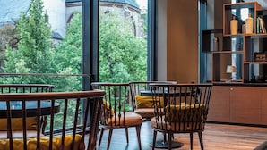 Um interior moderno com área de refeições, com cadeiras e mesa de madeira, espaço de bar com balcão e prateleiras, e uma janela ampla com vista para árvores e um edifício.