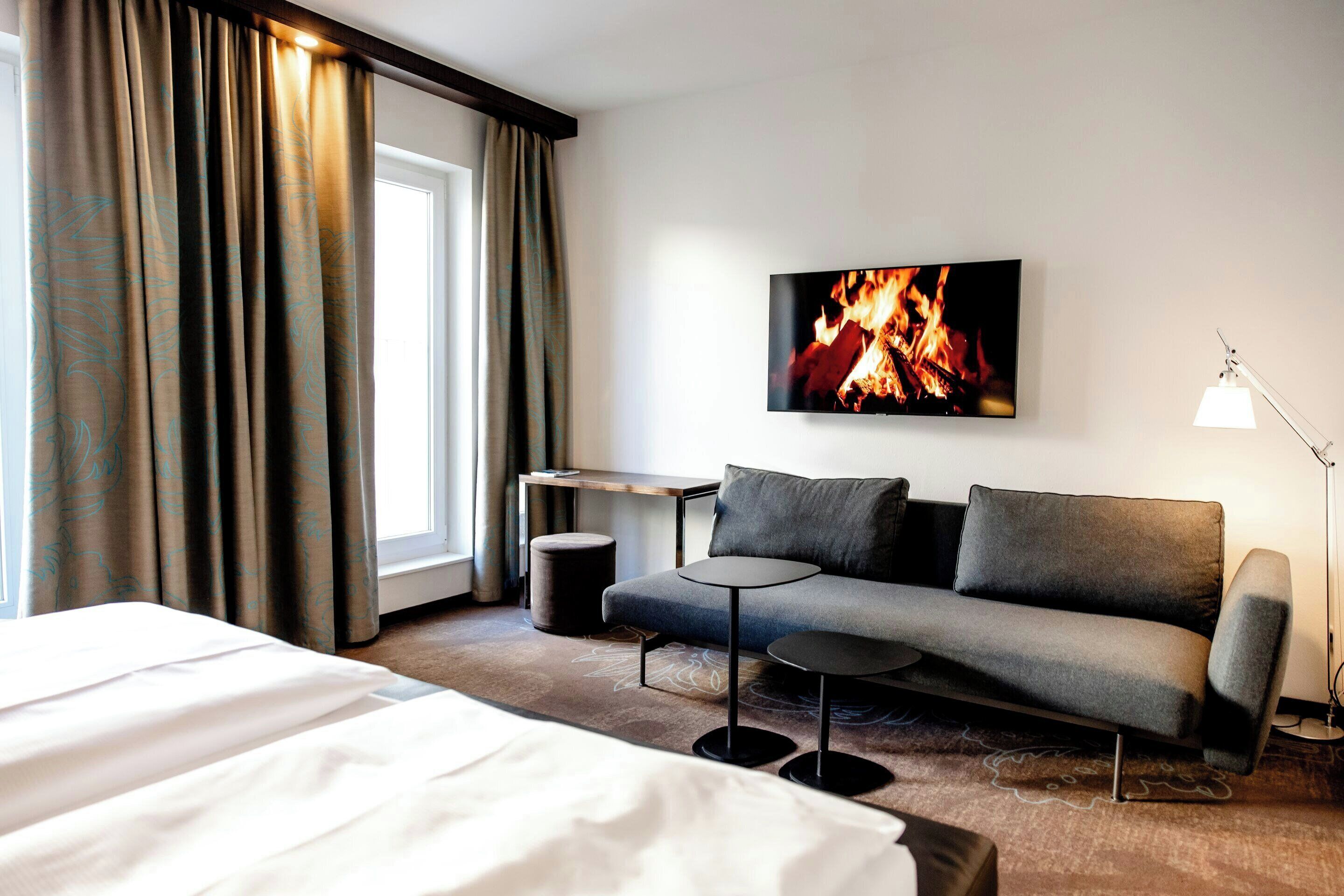 Ett hotellrum med en säng, en soffa, ett litet bord, en lampa och en tv som visar en eld.