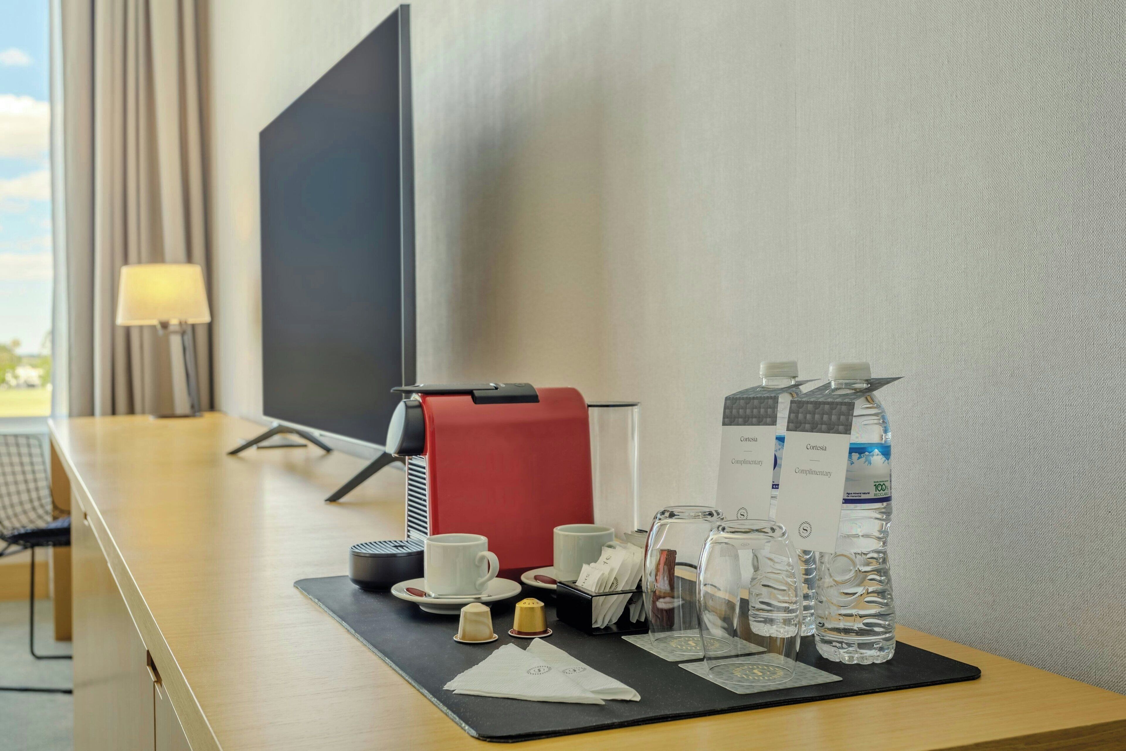 Habitación de hotel con escritorio, cafetera, tazas, botella de agua y jarra de vidrio.