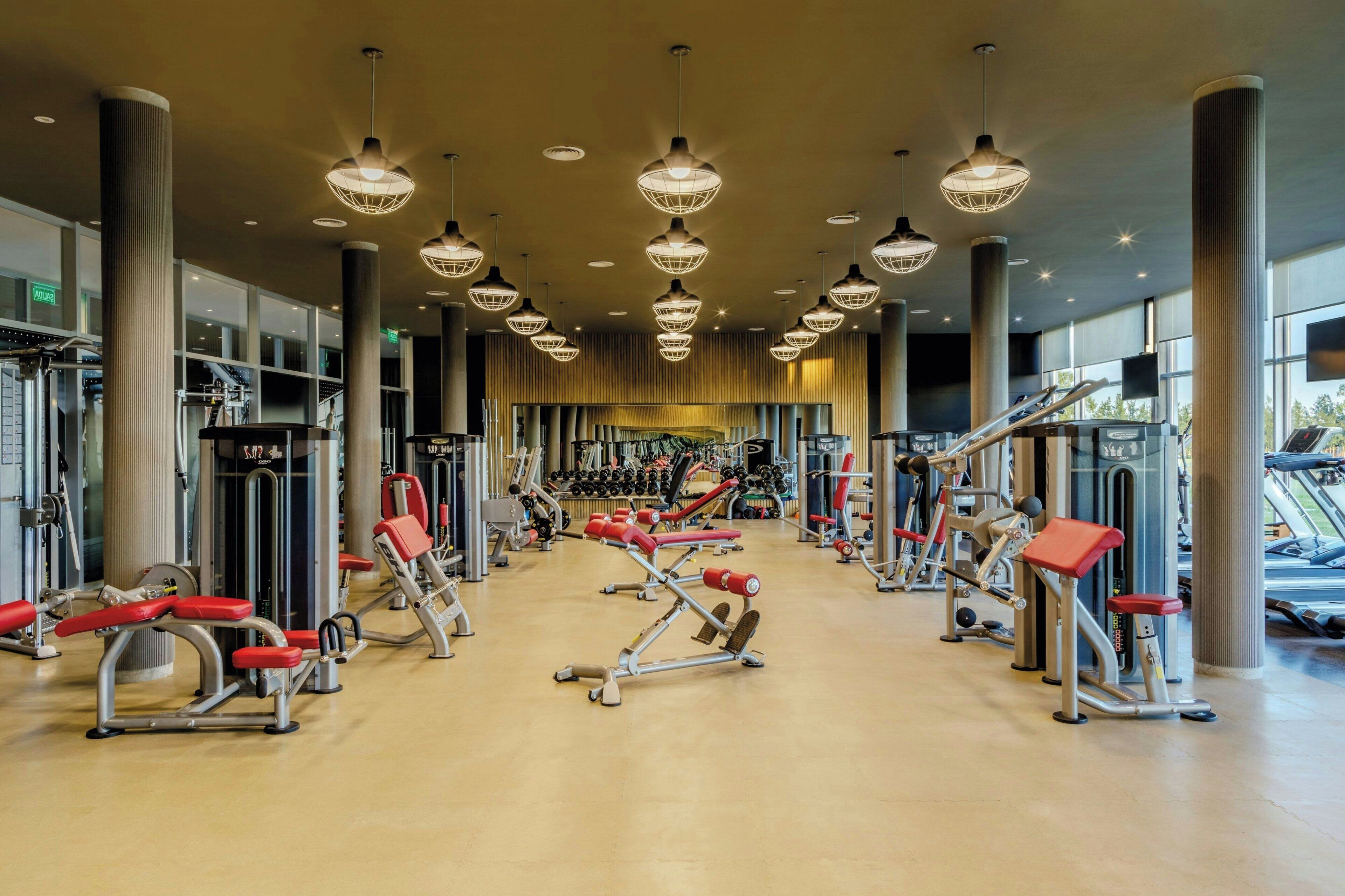 Un gimnasio amplio con diversos equipos de ejercicio, ventanas grandes y un diseño de techo moderno.