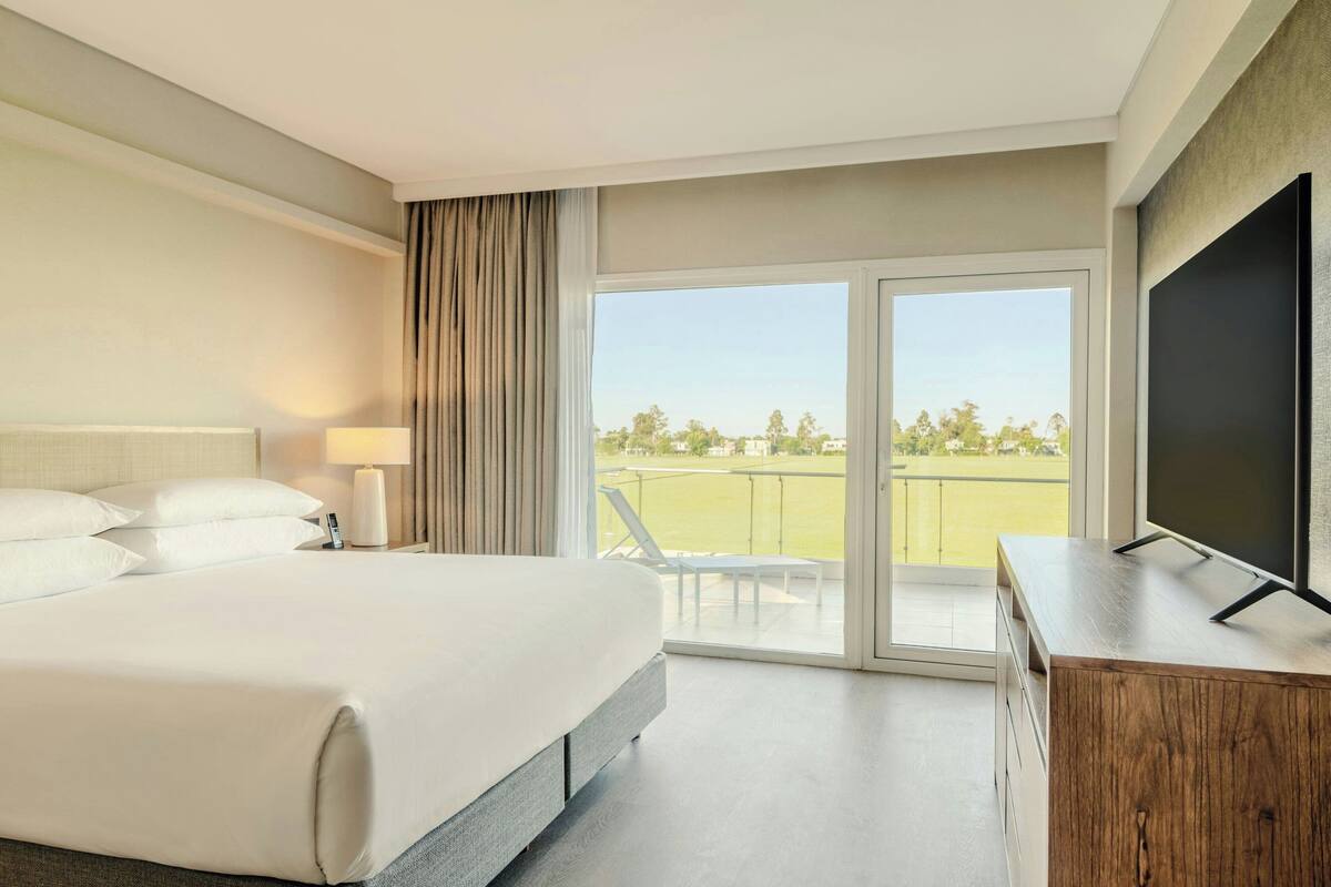 A hotel room with a bed, a TV, a desk, and a balcony with a view of a green field.