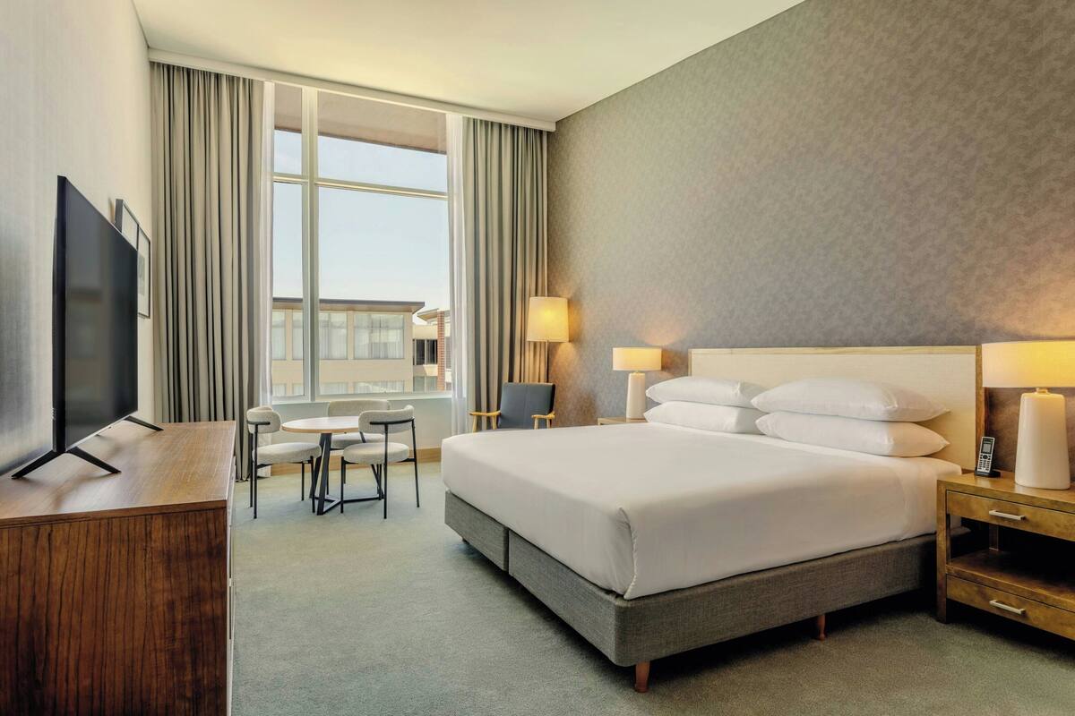 A hotel room with a large bed, bedside lamps, a desk with a flat-screen TV, a small table with chairs, and a large window with curtains.